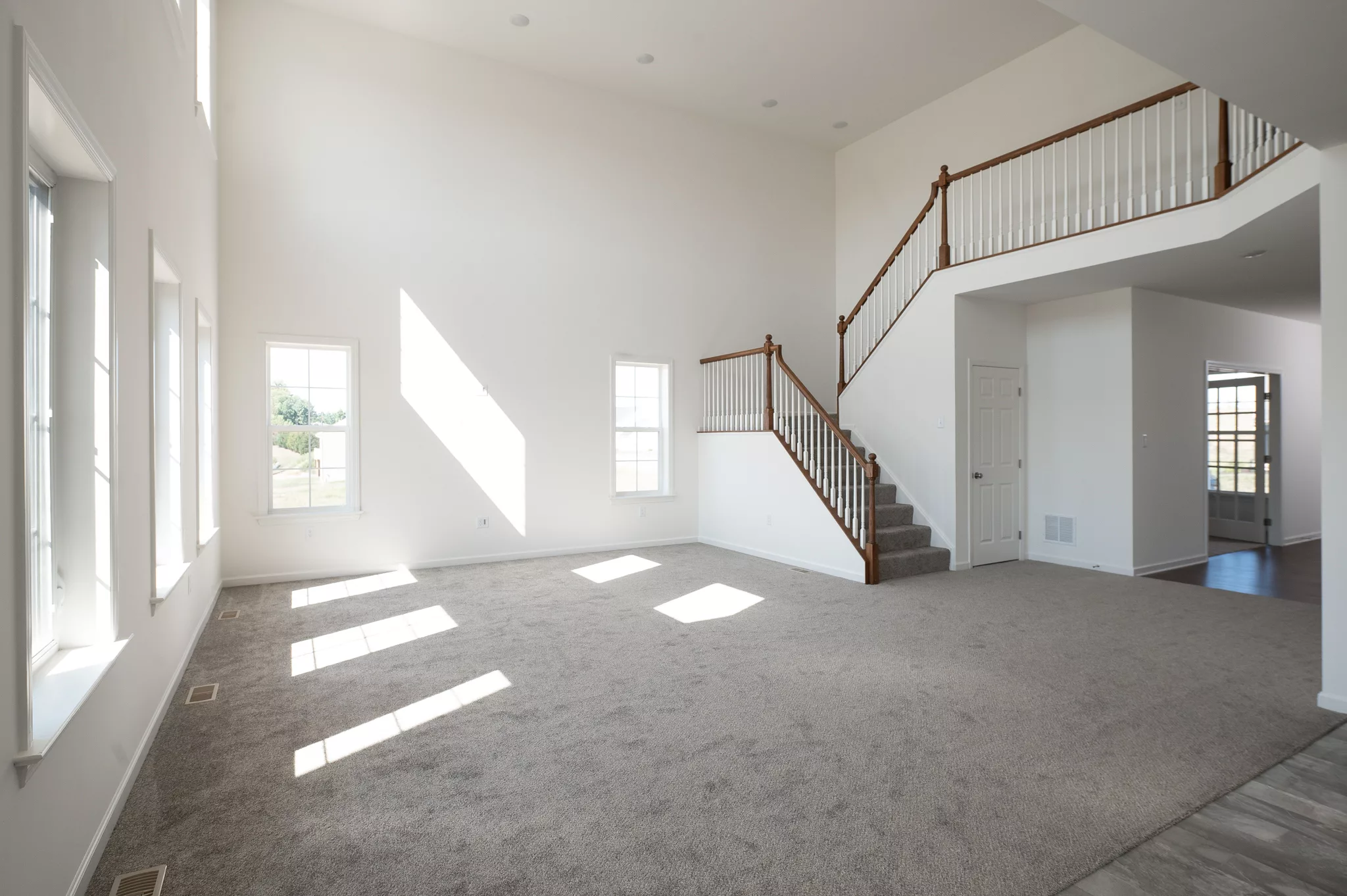 A spacious, bright living room in Lawrenceville with large windows, carpeted floor, white walls, and a staircase with wooden railings leading upstairs. Sunlight streams through the windows, creating patterns on the floor.