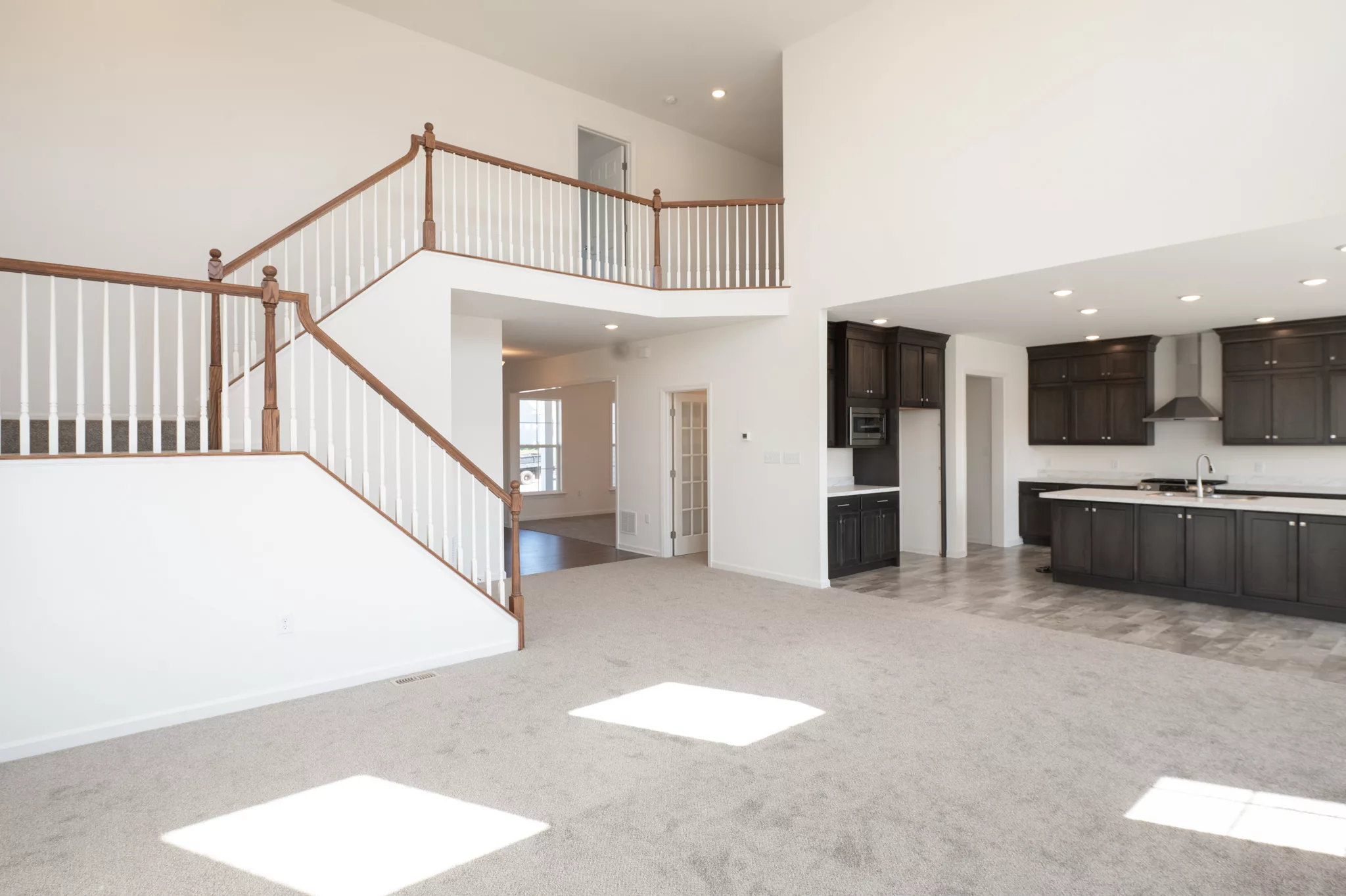 Spacious open-concept living area in Lawrenceville with beige carpet, large windows, a staircase with wooden railings, and a modern kitchen featuring dark cabinets and stainless steel appliances.