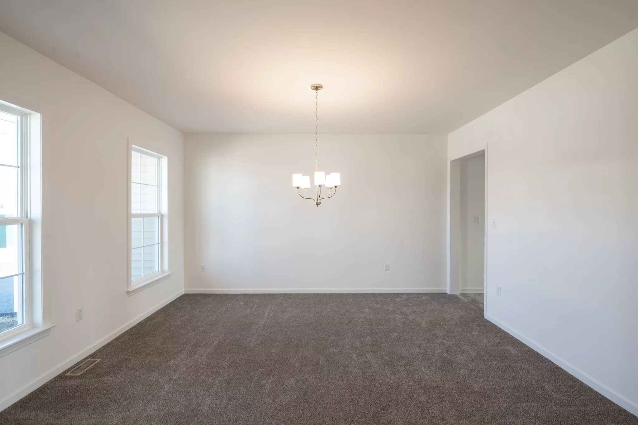 A bright, empty Lawrenceville room with white walls, two large windows, gray carpet, and a central ceiling light fixture with five bulbs hanging above.
