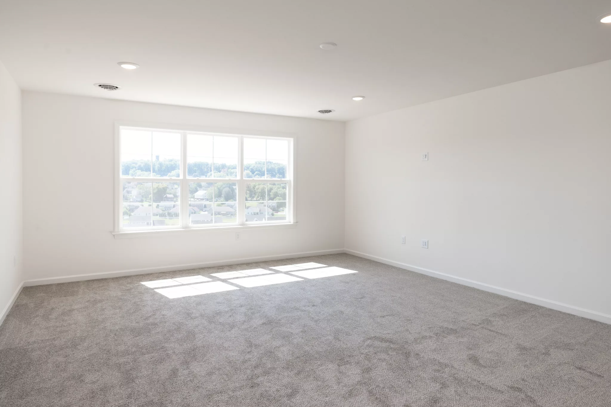 Bright, empty Lawrenceville room with light gray carpet, white walls, and a large window letting in natural sunlight. The window offers views of trees and distant houses. Recessed ceiling lights enhance the spacious, clean feel.