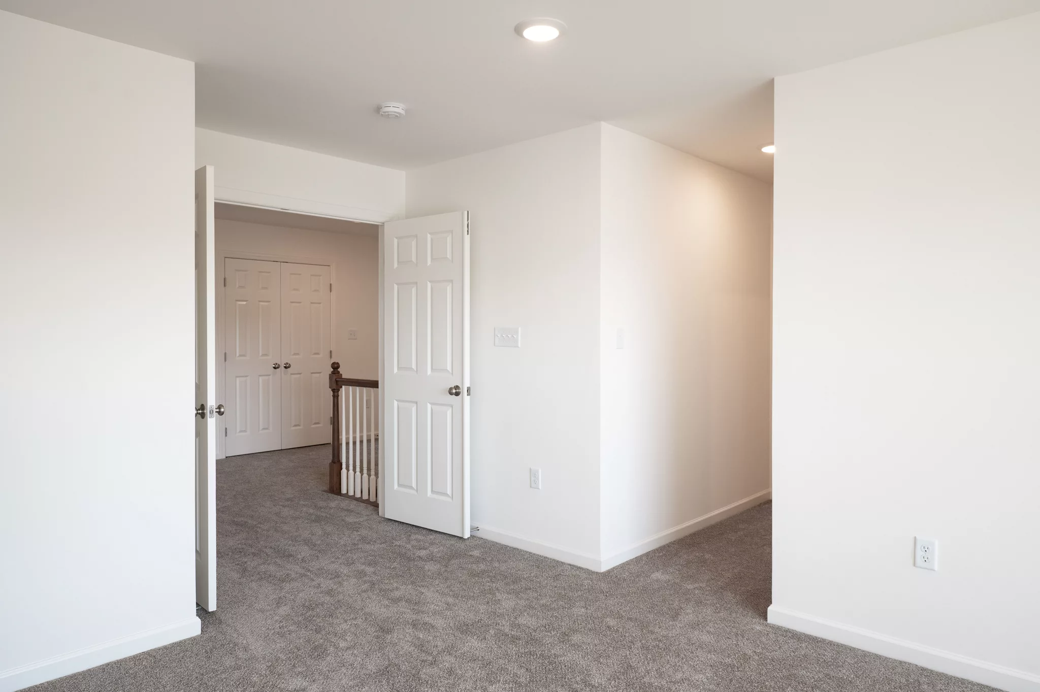 A bright, empty room with beige carpet, white walls, and recessed ceiling lights. Two white doors open to a hallway in this Lawrenceville home, revealing more closed doors and a staircase railing.