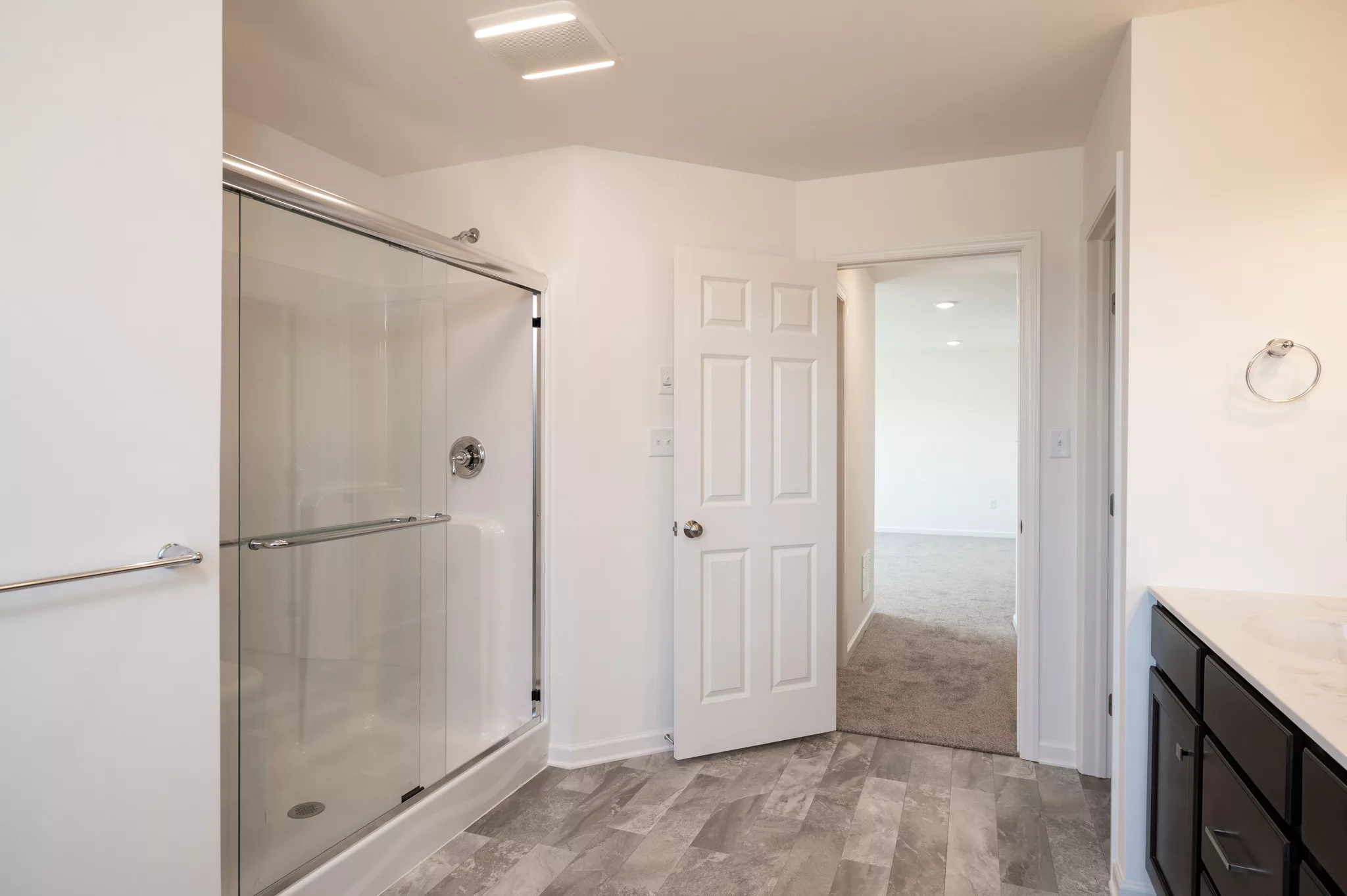 Modern Lawrenceville bathroom with a glass shower enclosure, a white door leading to a carpeted room, gray tile floor, and a black vanity with a white countertop and towel ring on the wall.