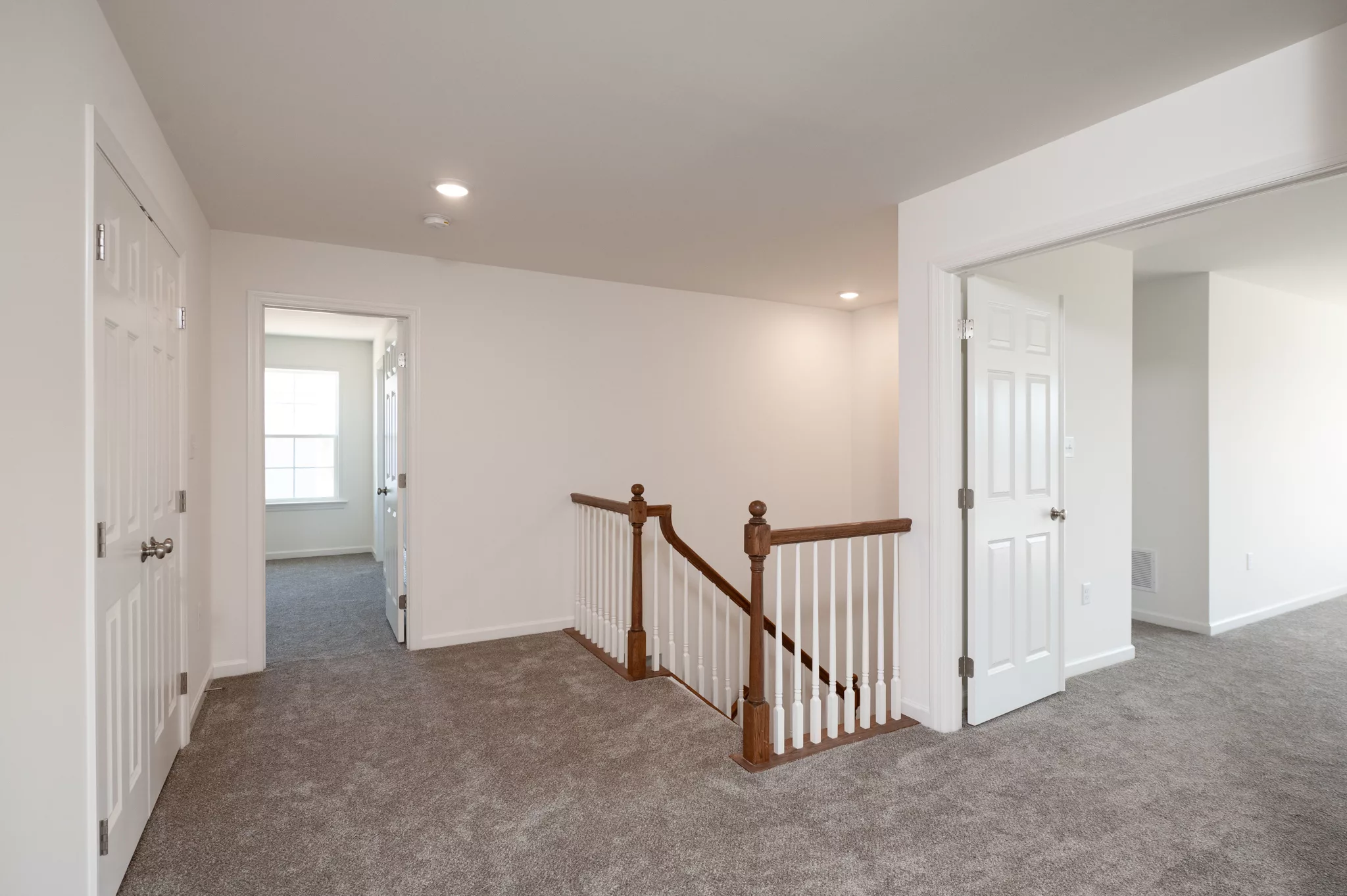 Bright upstairs hallway in this modern Lawrenceville home features beige carpet, white walls, a wooden handrail staircase, and open doors to other rooms. Natural light pours in from a window at the end of the hall.
