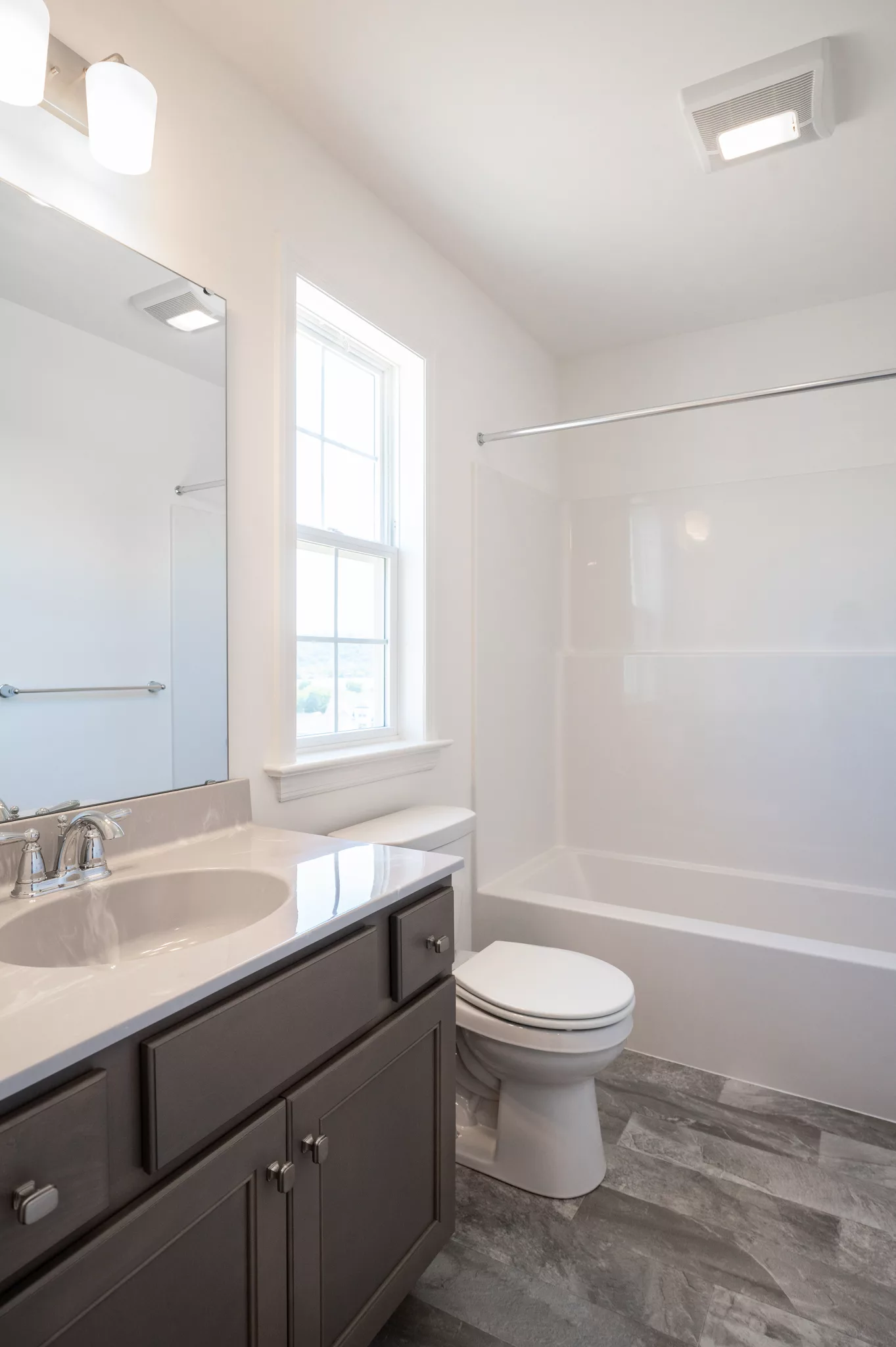 Modern Lawrenceville bathroom with a gray vanity, white sink, chrome faucet, large mirror, toilet, and white shower-tub combo. Sunlight streams through a window, brightening the space with gray tile flooring.