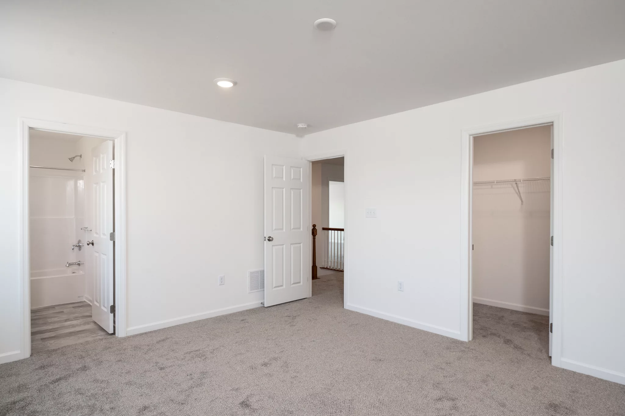 A bright, empty Lawrenceville bedroom with light-colored carpet, white walls, two open doors to a bathroom and walk-in closet, plus a third open door facing a hallway with a railing.
