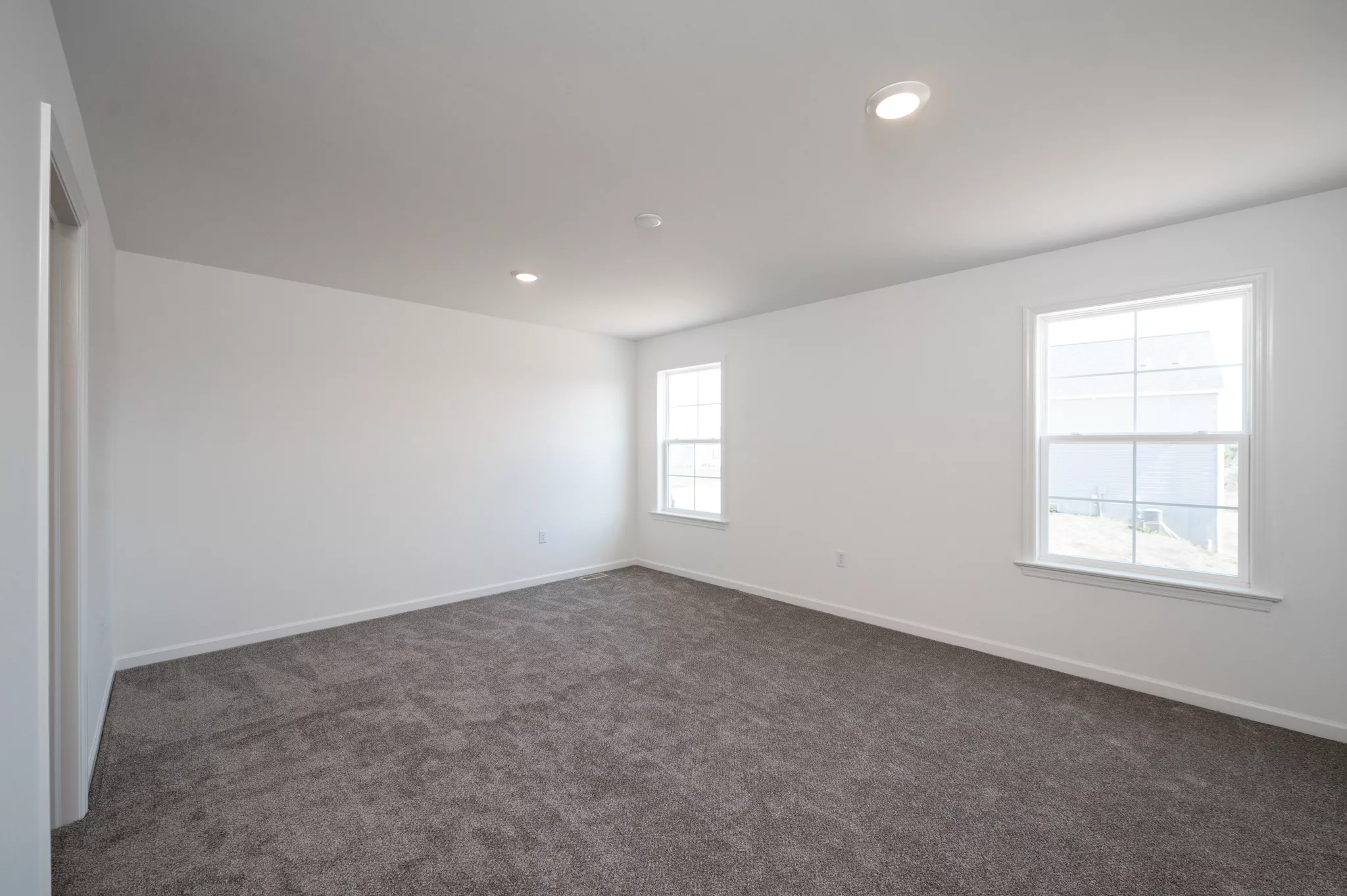 A bright, empty room in Lawrenceville with white walls, gray carpeted floor, two large windows letting in natural light, and recessed ceiling lights.