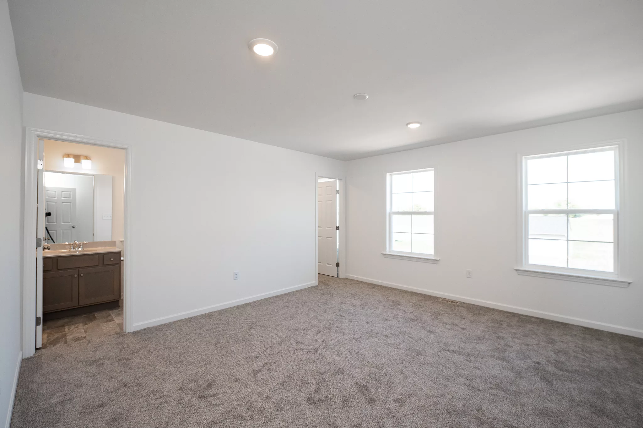 A spacious, empty room in Lawrenceville features light gray carpet, white walls, two large windows, and a doorway leading to a bathroom with double vanity and mirror. Ceiling lights brighten the airy space.