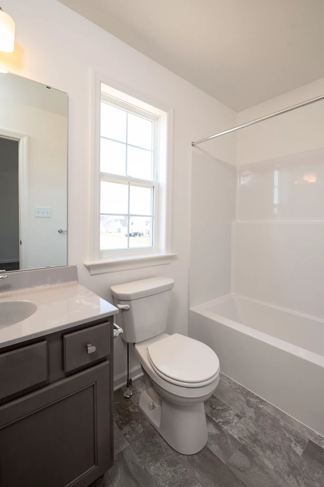 A bright Lawrenceville bathroom with a white bathtub and shower, toilet, vanity with a sink and gray cabinet, a large mirror, and a window letting in natural light. The floor is gray and the walls are white.