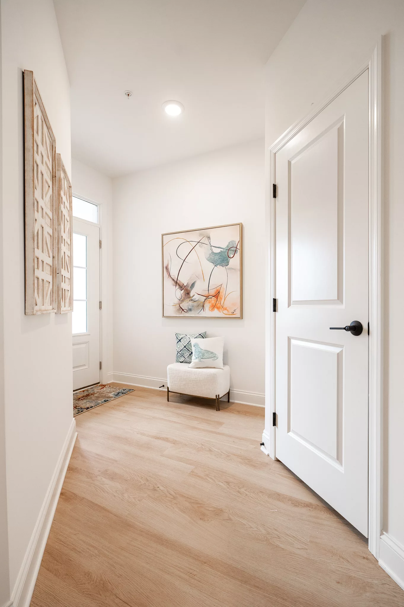 A bright hallway with light wood floors and white walls, featuring abstract art, a small upholstered chair with pillows, and a windowed door letting in natural light.