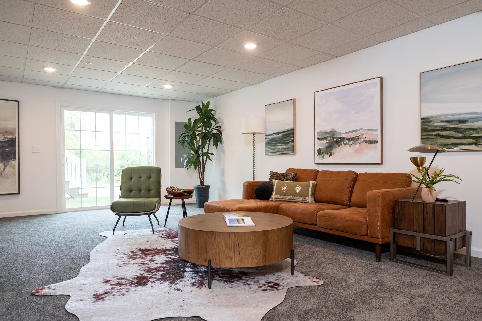 Modern living room with a brown sofa, green chair, round wooden coffee table on a white cowhide rug, wall art, large plant, and a sliding glass door letting in natural light.