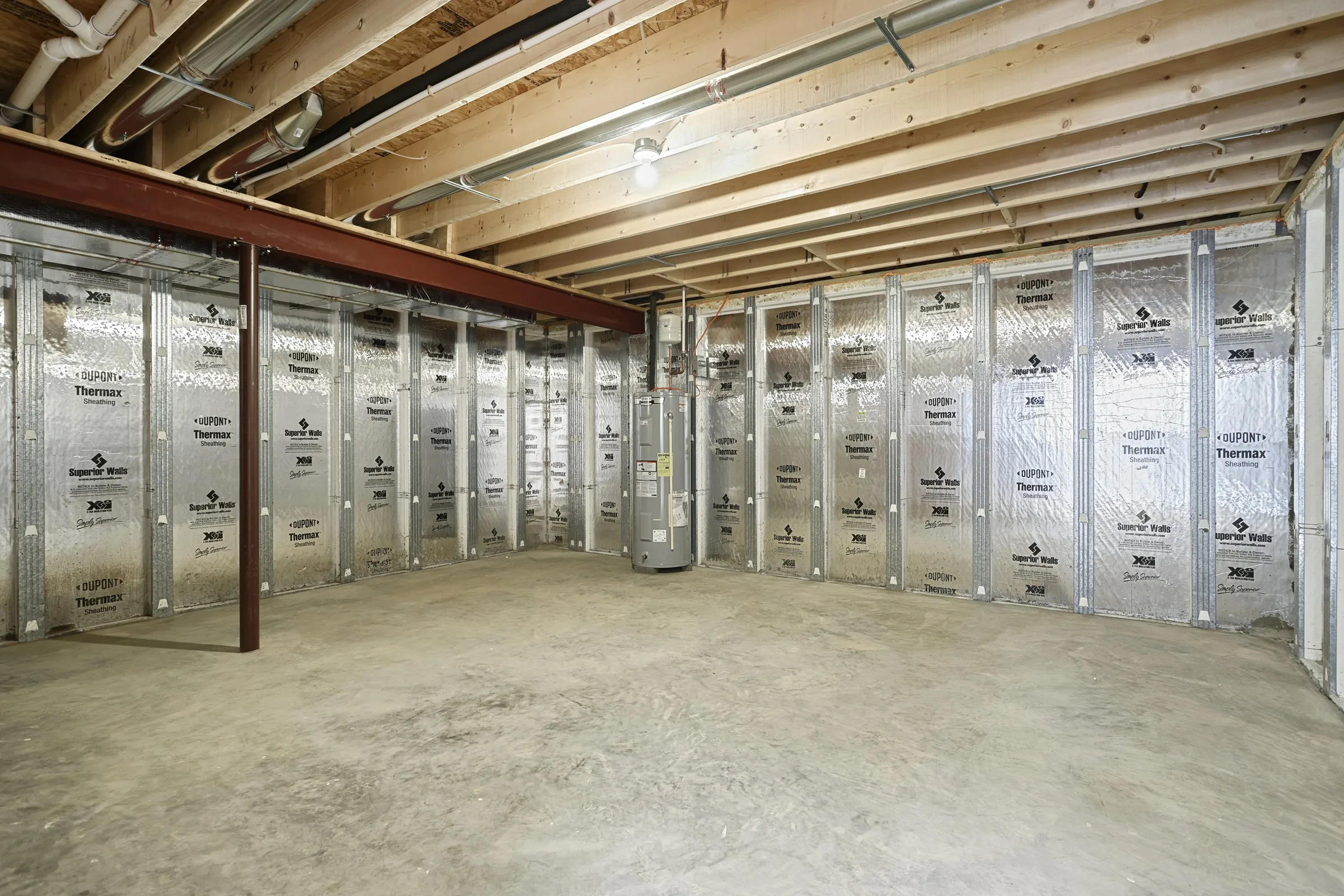 Unfinished basement with exposed wooden beams and insulated walls, concrete floor, visible pipes, and a hot water heater in the corner under a ceiling light.