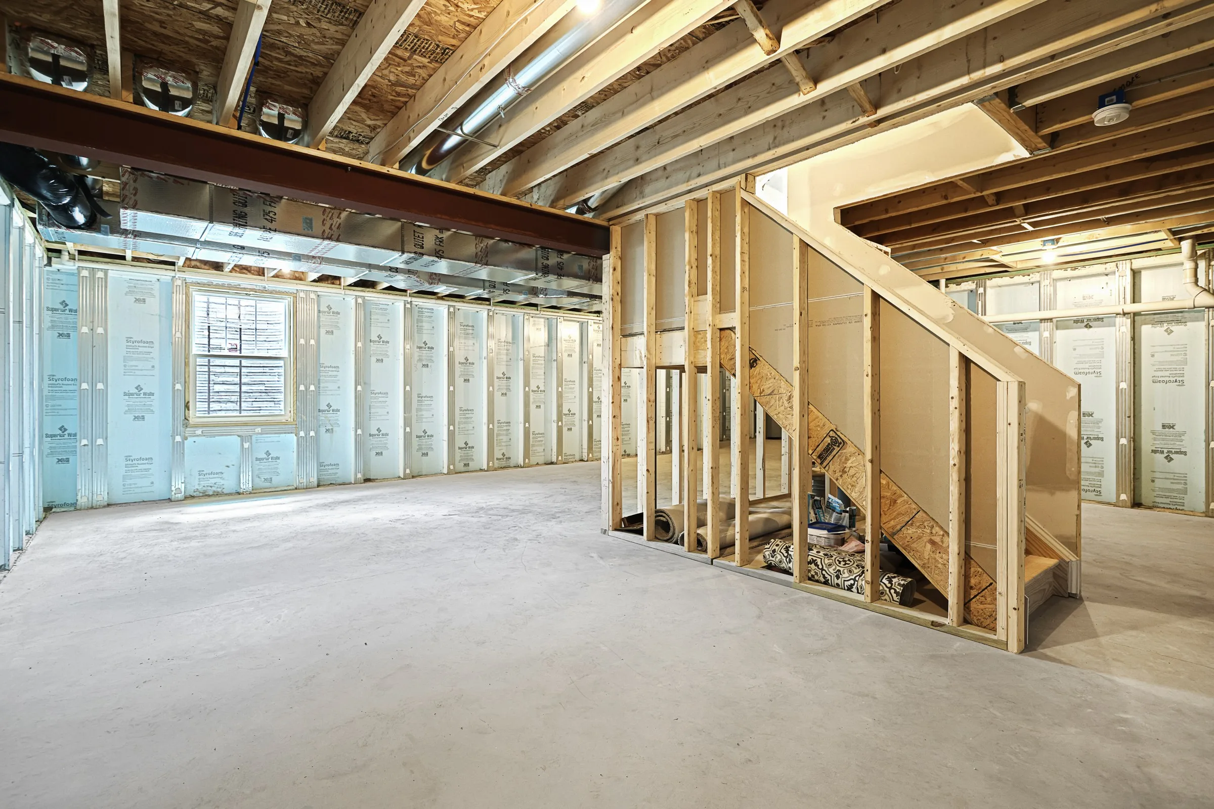 Partially finished basement with exposed wooden beams and insulation, concrete floor, and an unfinished staircase. Light enters through a window on the left wall. The space appears open and under construction.