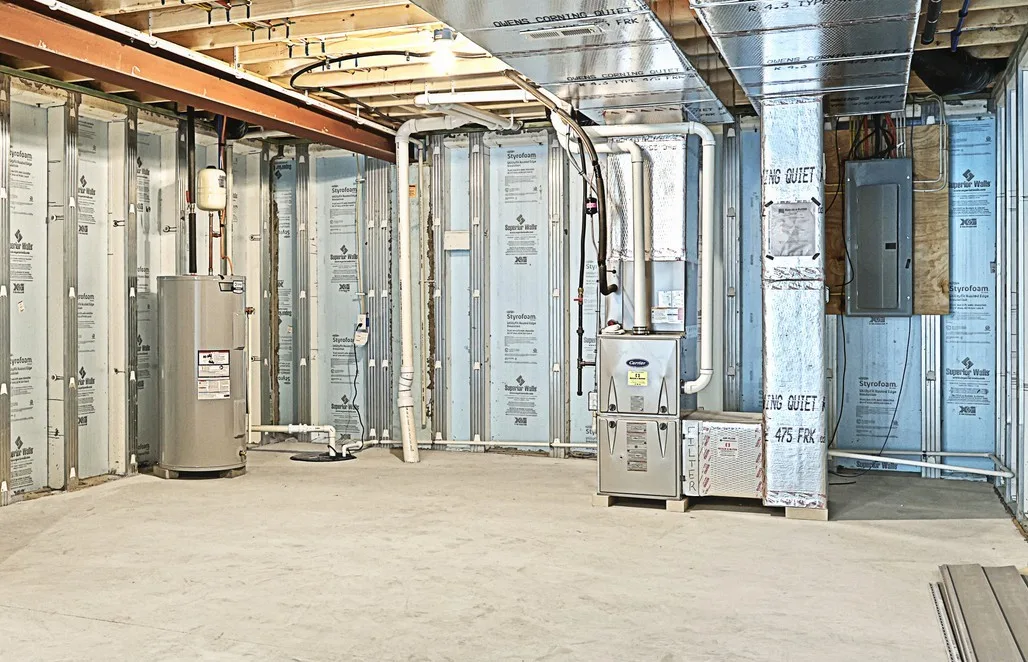 Unfinished basement with exposed insulation, pipes, and electrical wiring. A water heater and furnace are installed along the walls, and there is a concrete floor with no finished ceiling or walls.