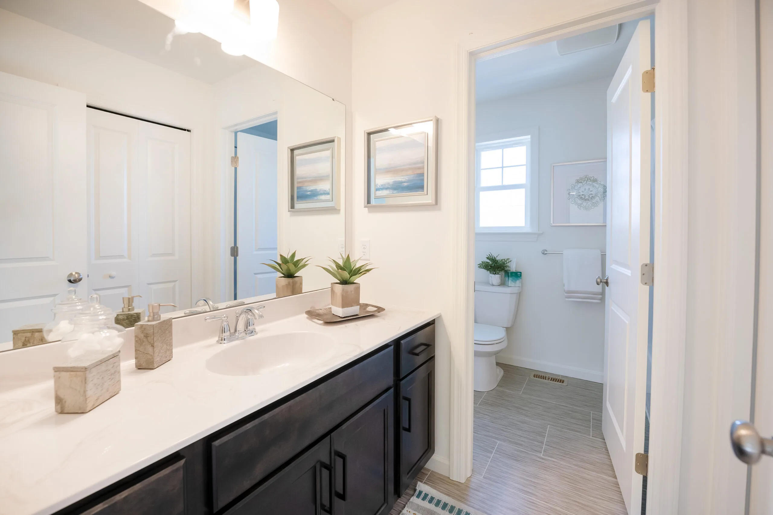 A bright, modern bathroom with a large mirror, double sink vanity with dark cabinets, two potted plants, and decorative items. A door leads to a toilet area with a window and towel rack.