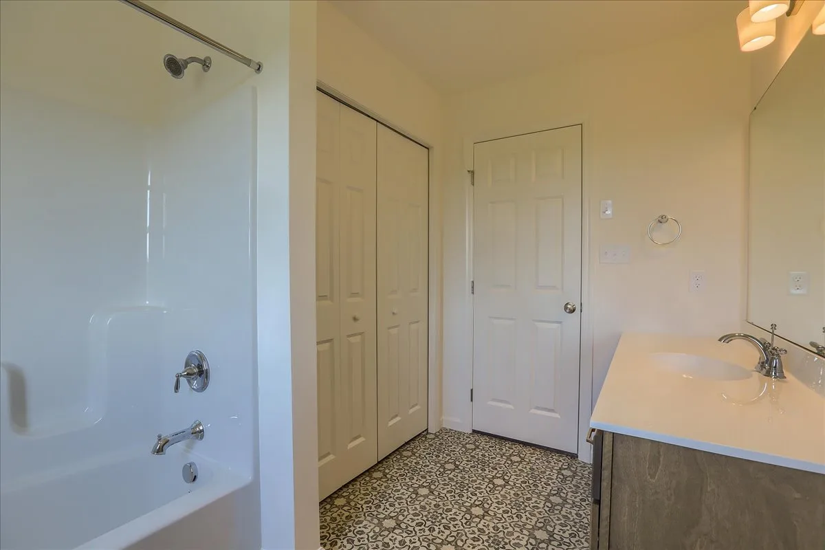 A bathroom with a white bathtub-shower combo, a vanity with a sink and mirror, patterned tile flooring, and two white closet doors. The walls are light-colored, and the lighting is soft and warm.