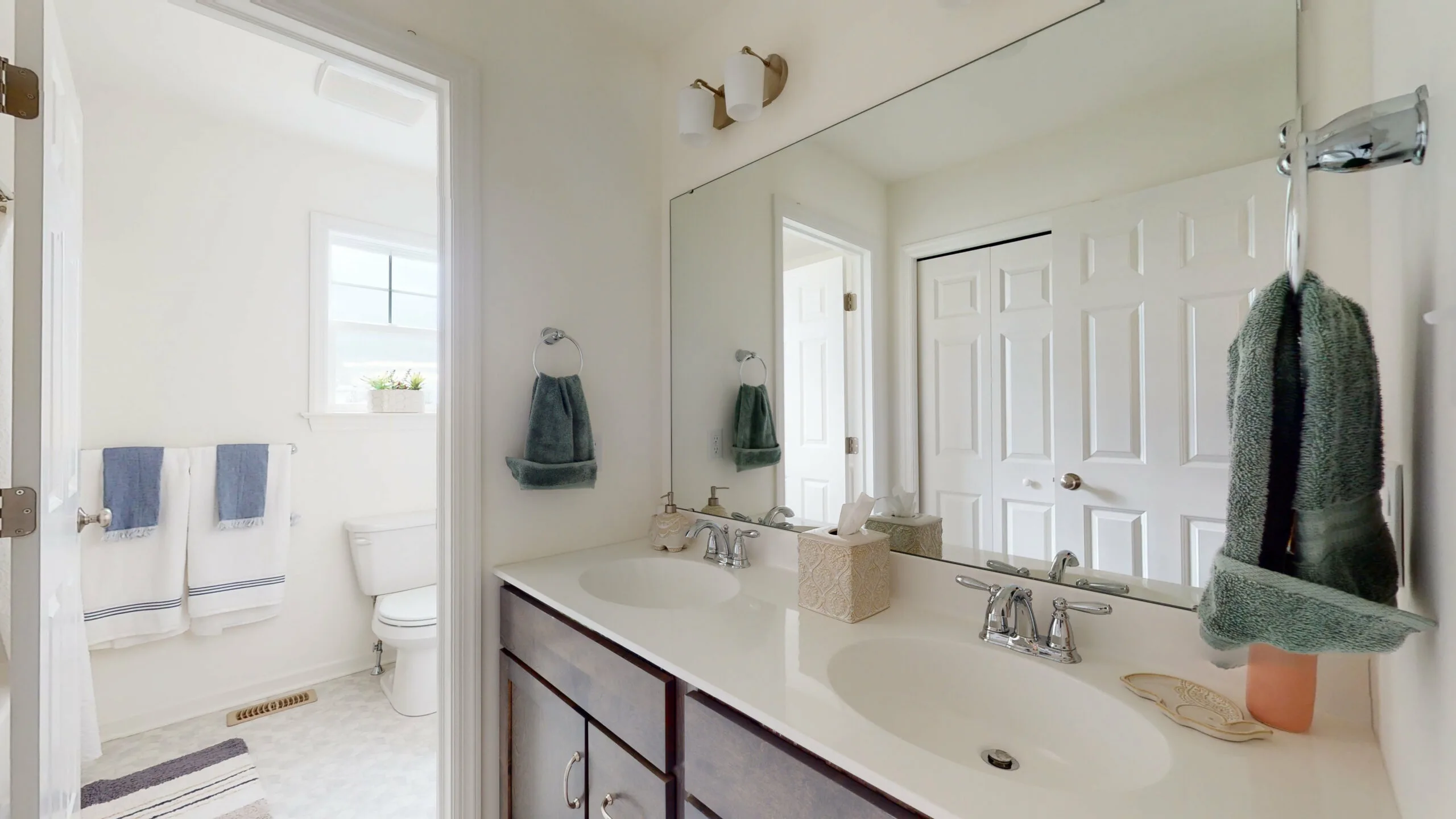 A bright, modern bathroom with a double sink vanity, large mirror, and dark wood cabinets. A white door leads to a separate toilet and shower area with a window and towel racks on the wall.