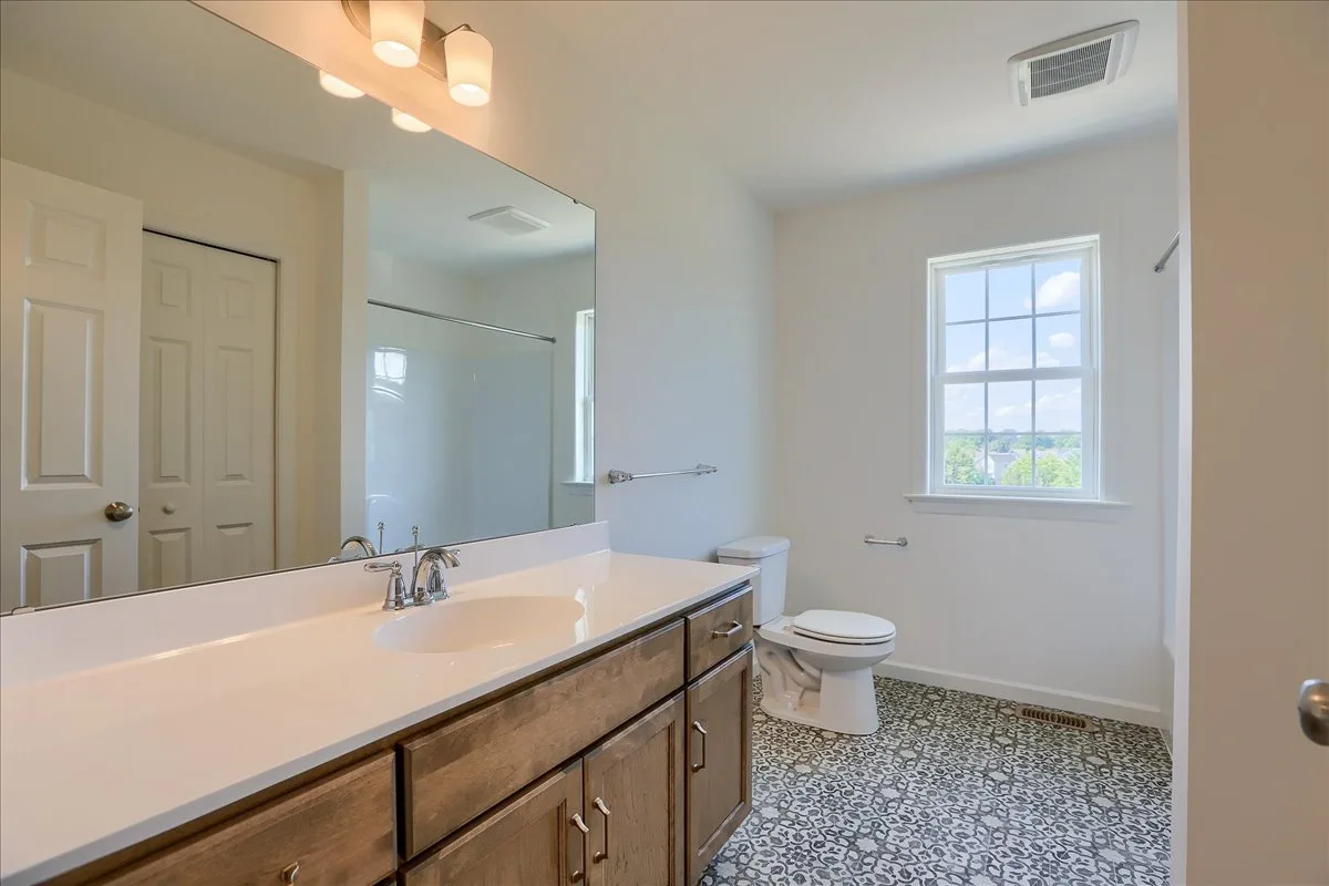 Bright bathroom with a large mirror, wooden vanity with sink, patterned tile floor, toilet, window with outdoor view, and a shower in the background.