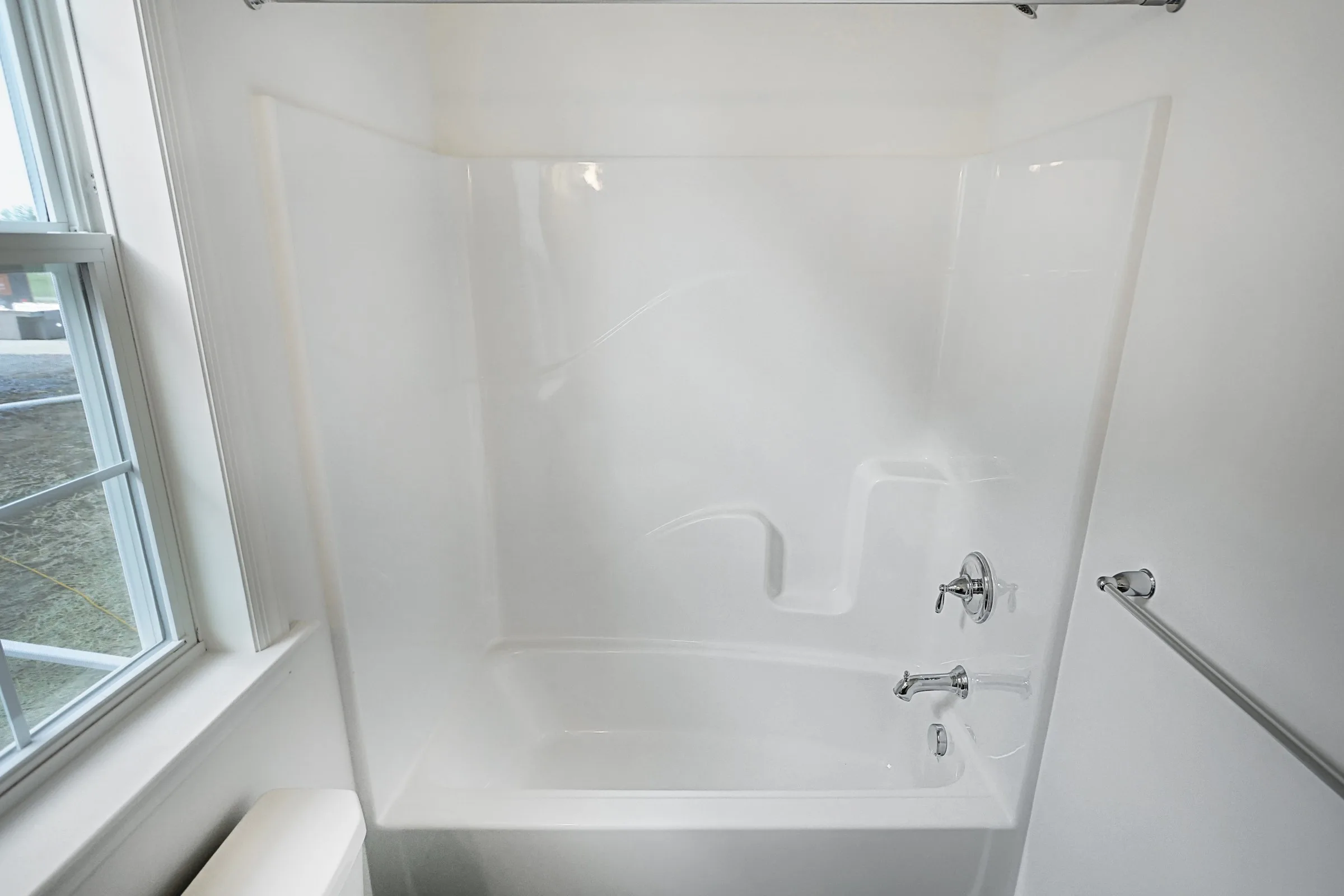 A clean, white bathtub with a built-in shower and chrome fixtures is shown in a bright bathroom with a window to the left and a towel bar on the right wall.