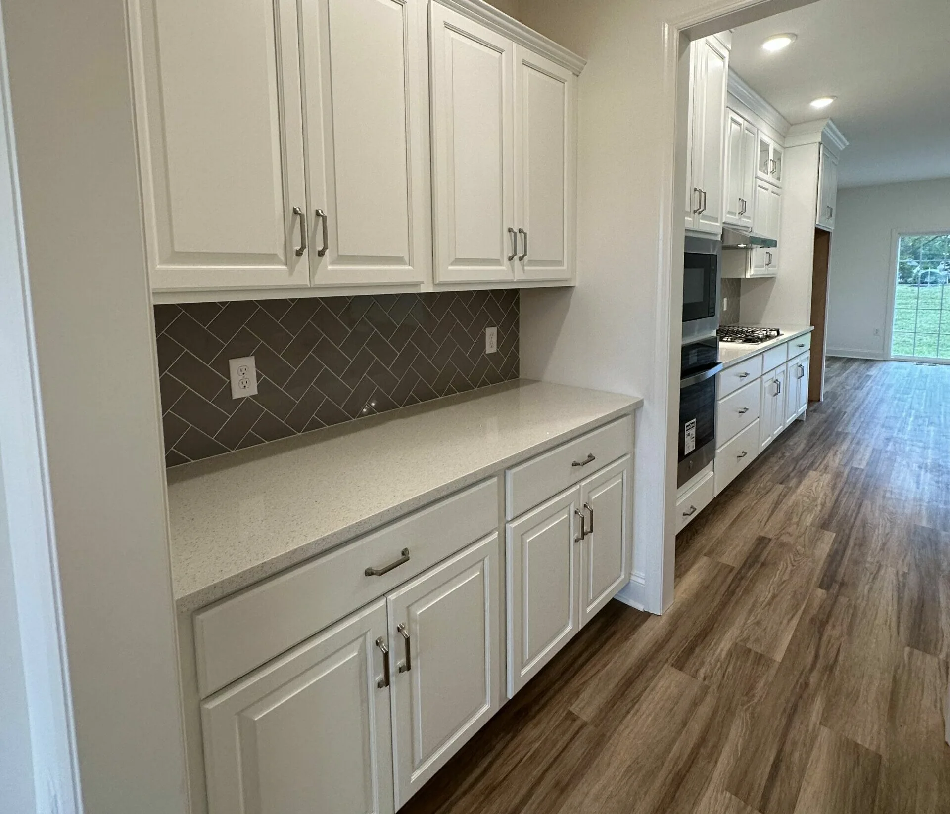 Modern kitchen with white cabinets, light countertops, brown herringbone tile backsplash, and wood-look flooring. Built-in oven and stove are visible, with a view towards a bright open living area and large window.