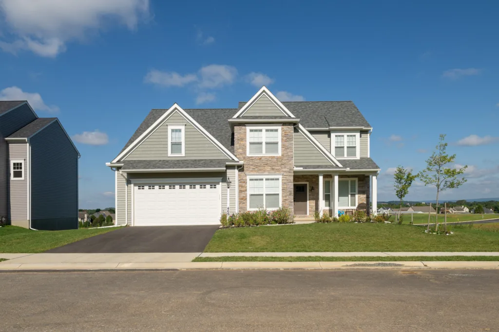 A modern two-story suburban house with light gray siding, stone accents, a two-car garage, and a well-kept lawn under a clear blue sky.