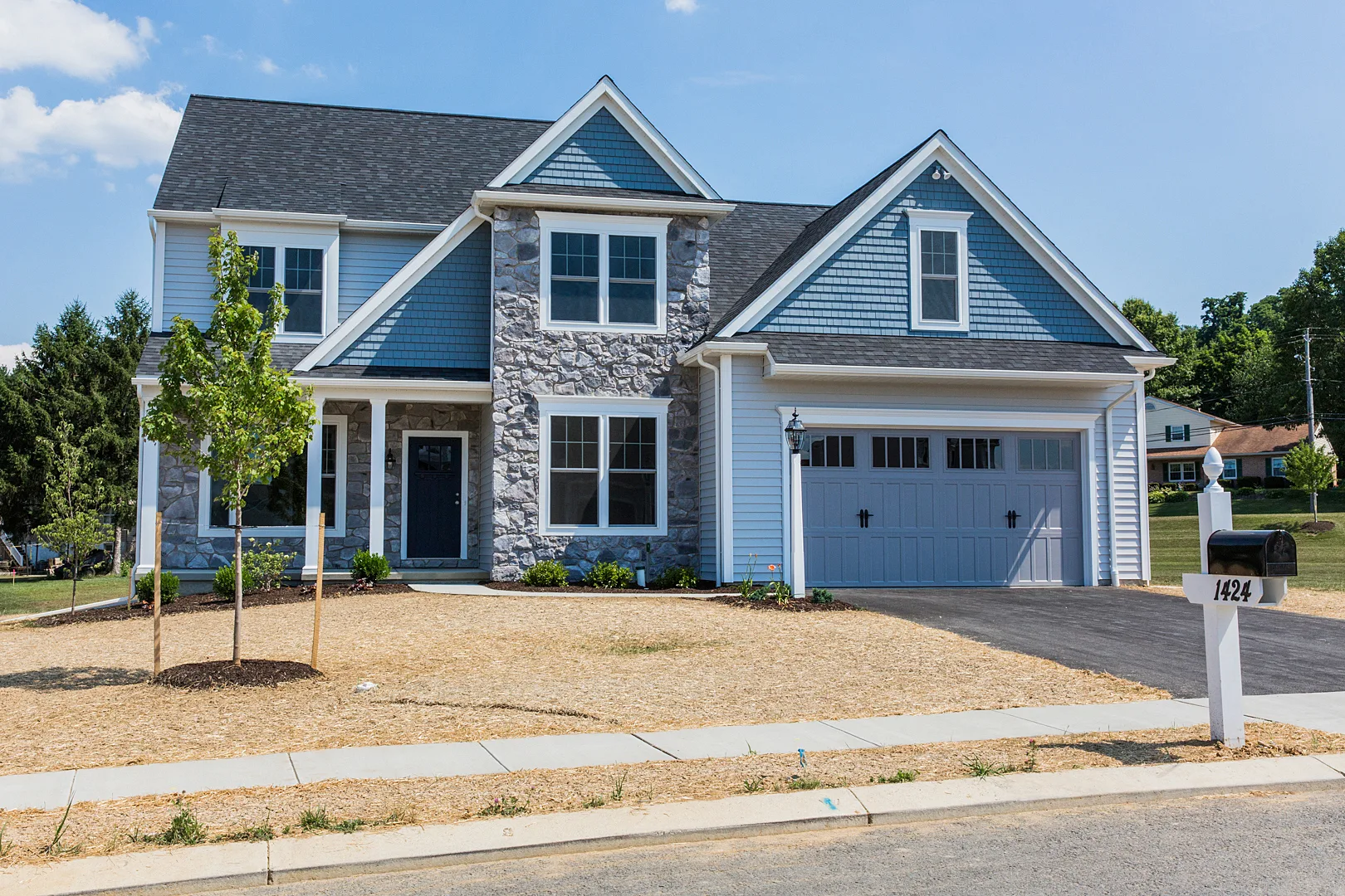 A two-story suburban house with blue-gray siding, stone accents, and a double garage. The front yard is freshly landscaped with young trees, mulch, and a white mailbox labeled 1424.