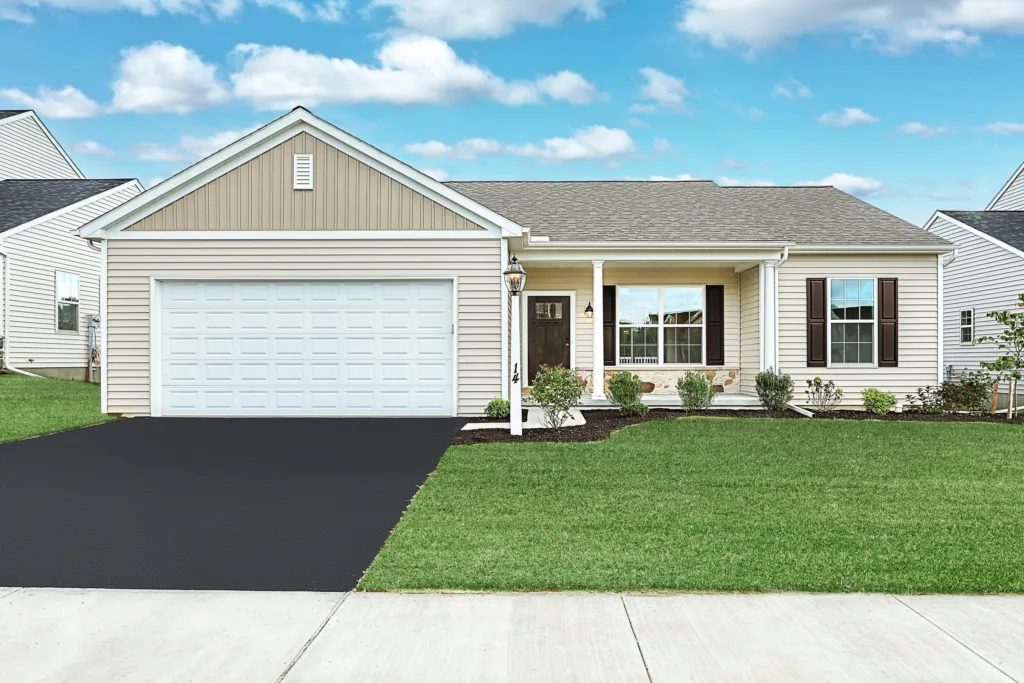 Single-story suburban house with beige siding, a two-car garage, front porch, and neatly landscaped front yard with green grass and bushes under a blue sky with scattered clouds.