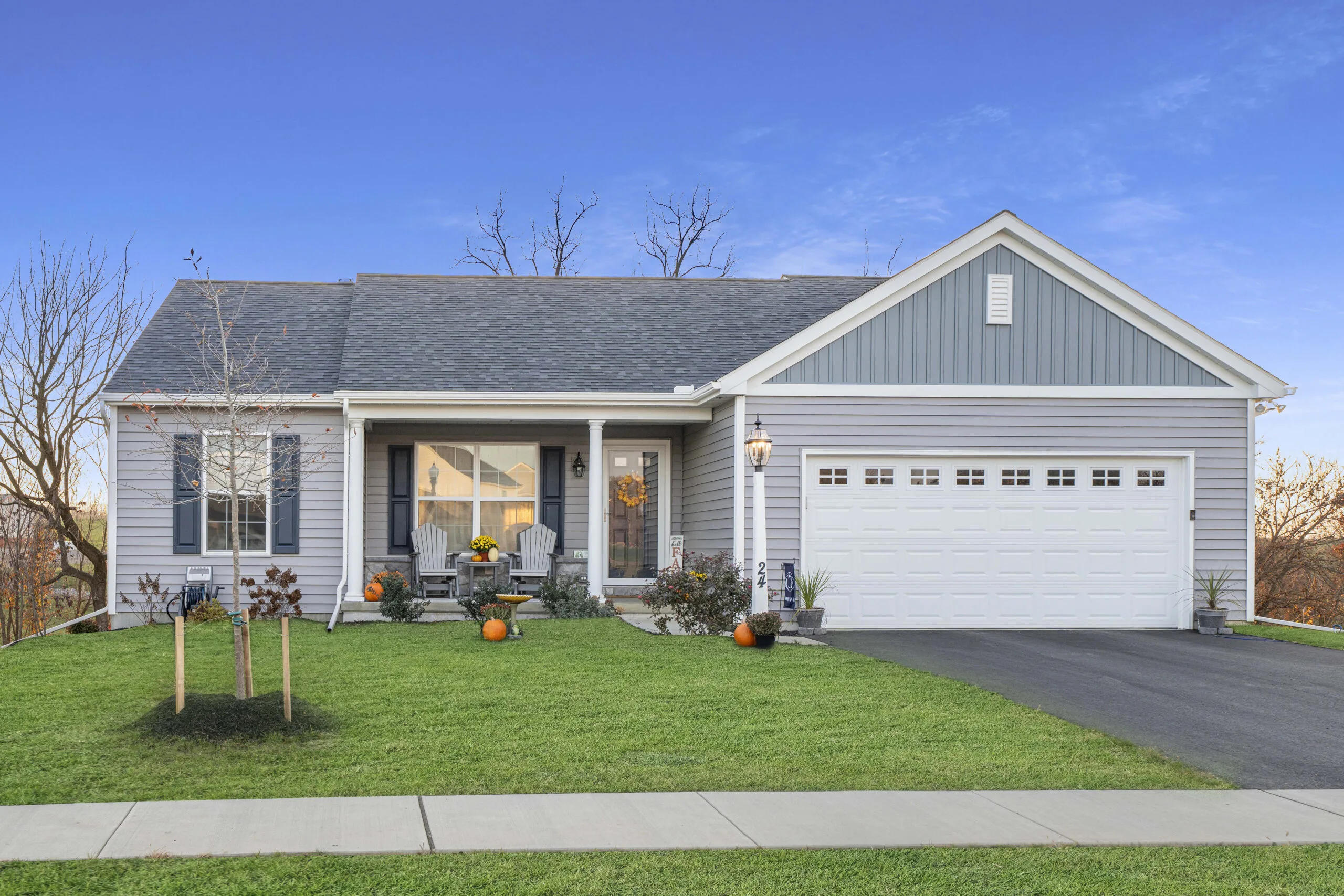 Single-story gray house with white trim, a front porch decorated with pumpkins and autumn flowers, attached two-car garage, and a well-kept green lawn under a clear blue sky.