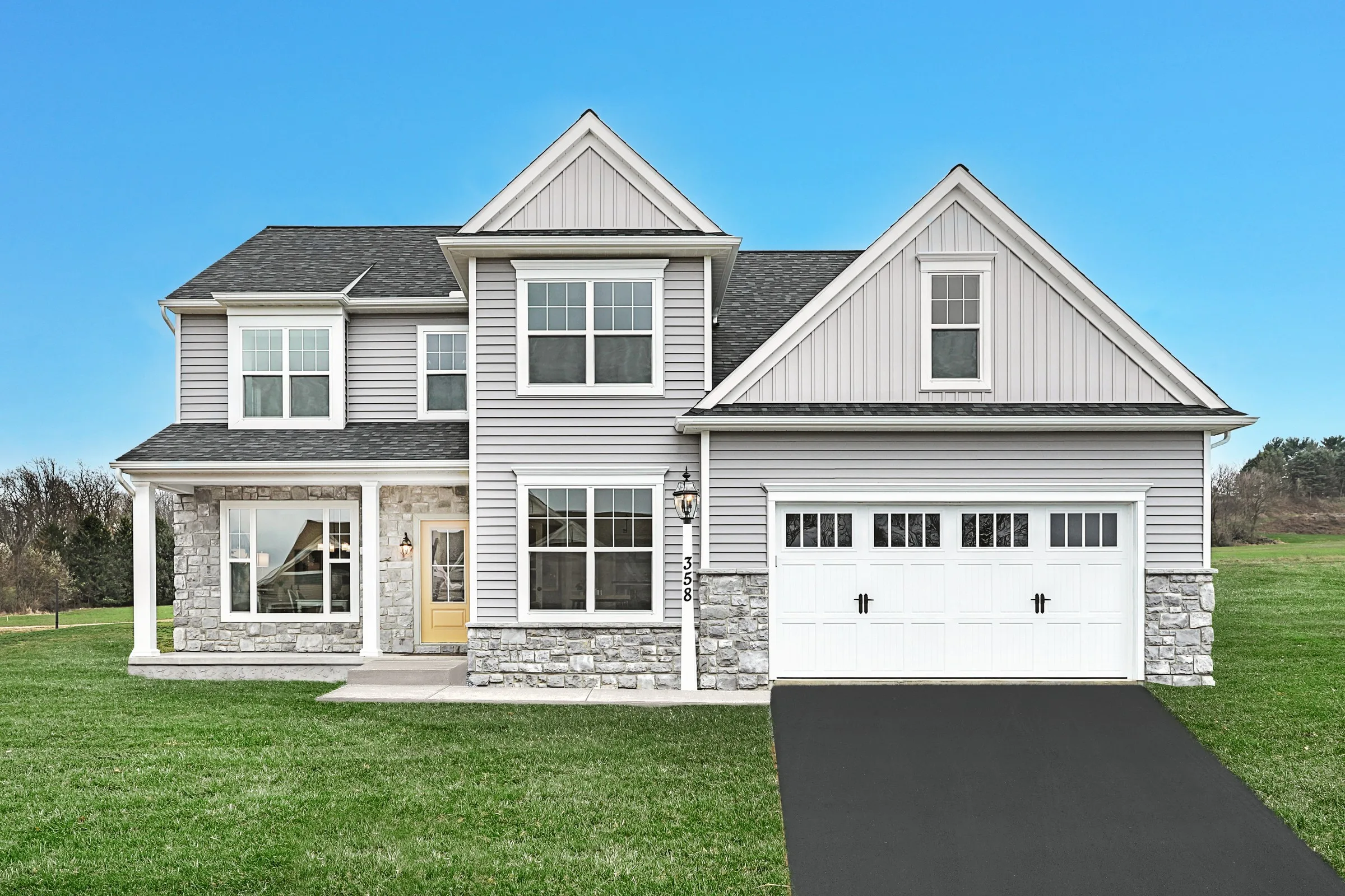 A modern two-story house with gray siding, stone accents, white trim, and a double garage. The home has large windows, a covered porch, and sits on a green lawn against a clear blue sky.