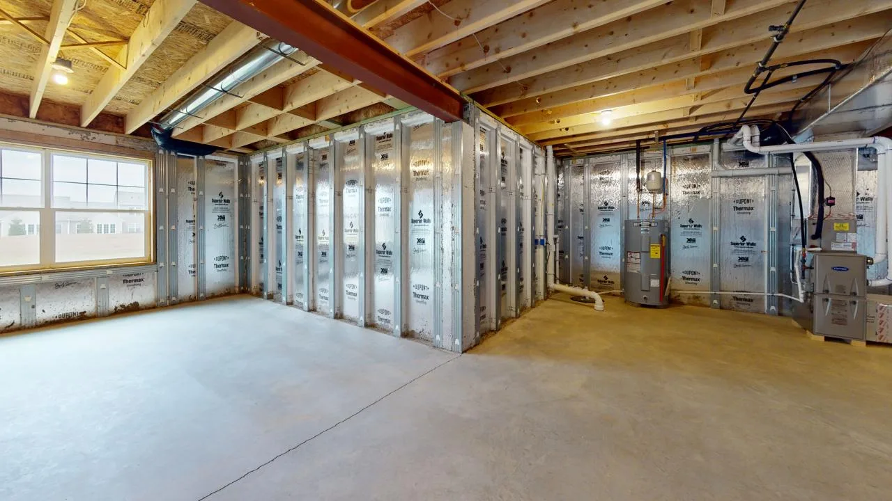 An unfinished basement with exposed beams, framed walls, and concrete flooring, featuring insulation on the walls and a water heater in the corner.