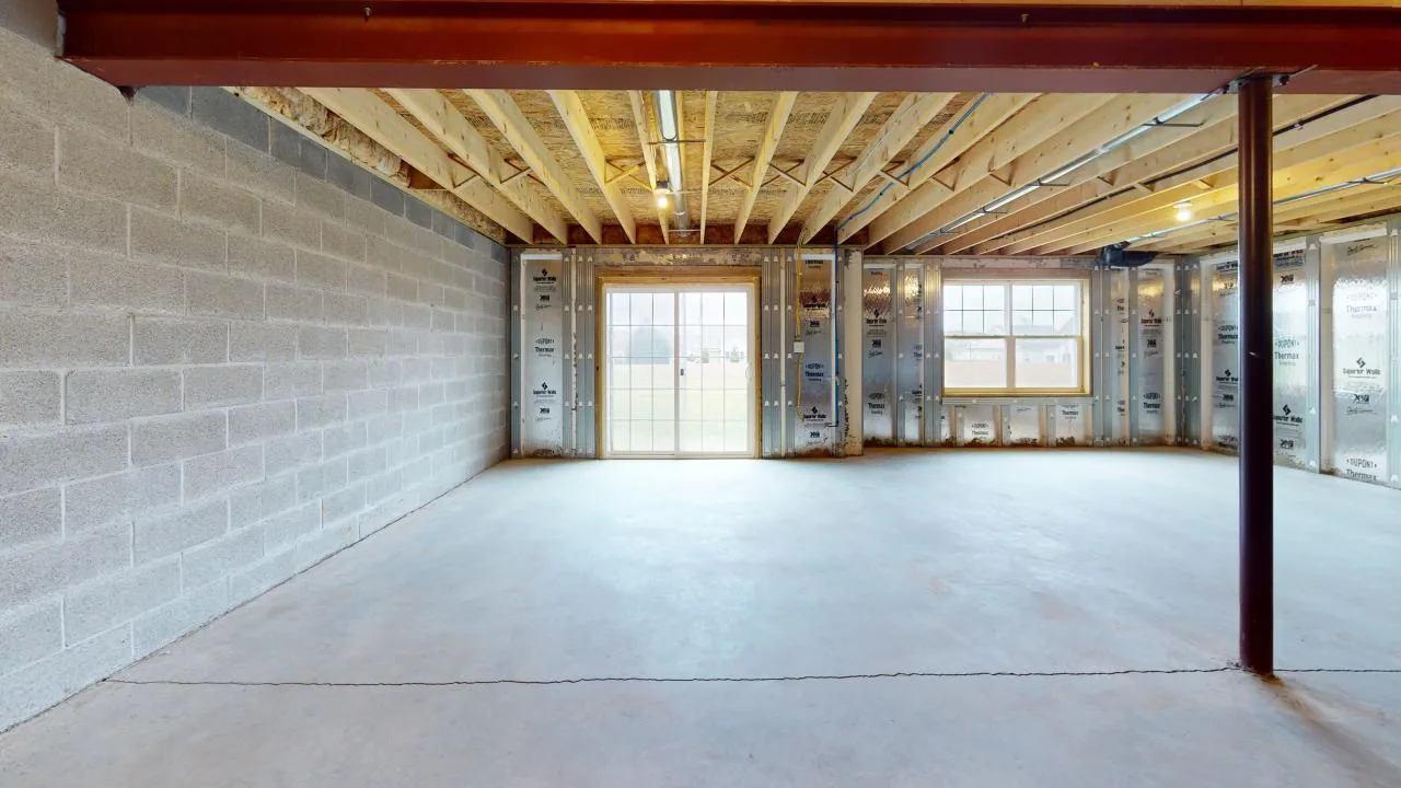 An unfinished basement with concrete floor, exposed beams, and partially insulated walls, featuring large windows and a door leading outside.
