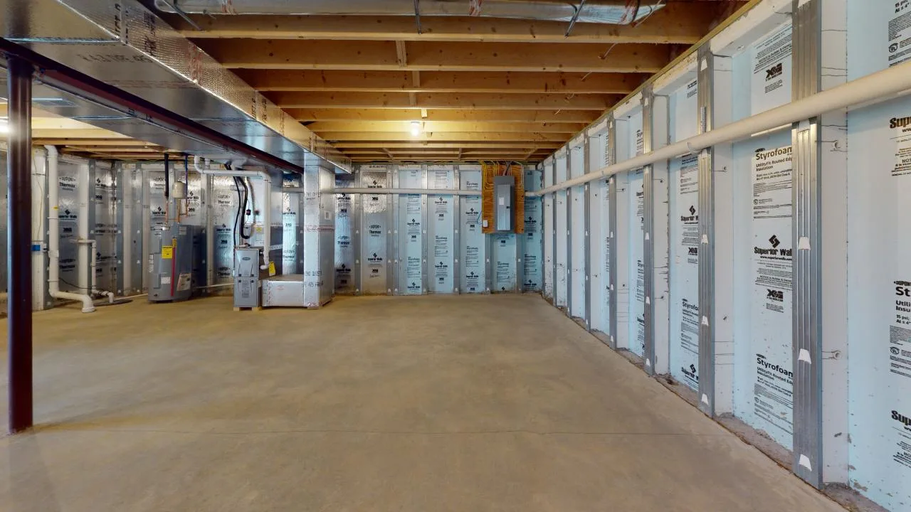 A spacious basement area with insulation on the walls and exposed wooden beams overhead, featuring concrete flooring and utility equipment.