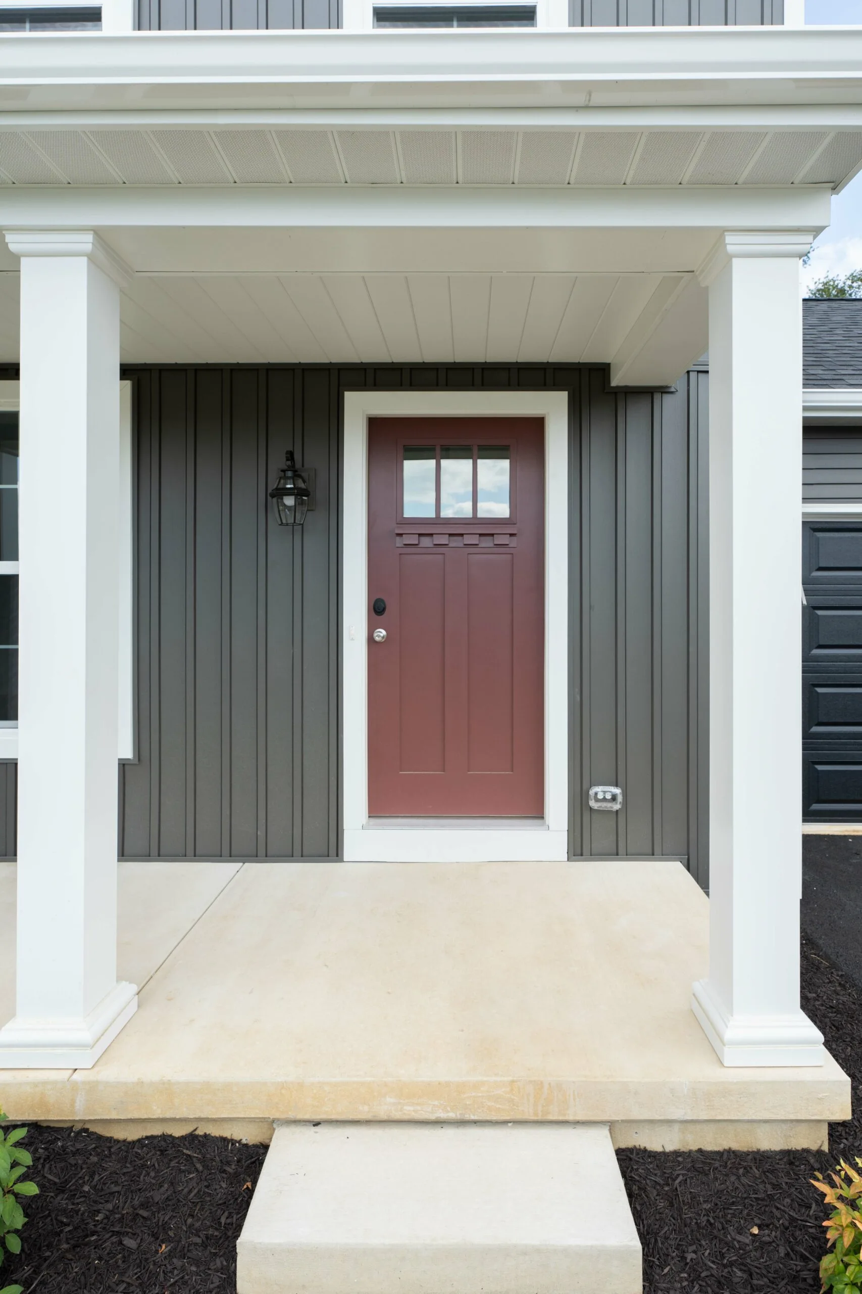 A modern front porch with white columns, a red door with square window panes at the top, gray vertical siding, a black wall lantern, and a concrete step leading to the entrance.