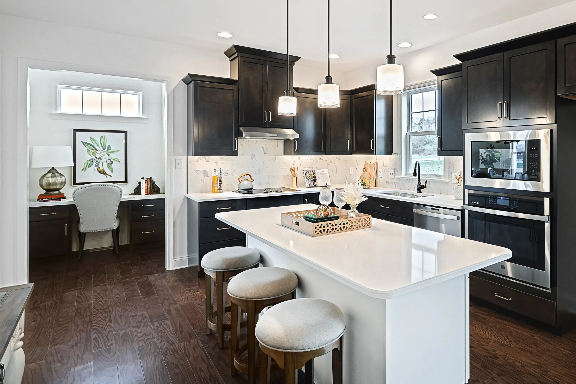 Modern kitchen with dark wood cabinets, white countertops, a center island with three round stools, pendant lights, built-in appliances, and an adjacent small office nook with a desk and chair.