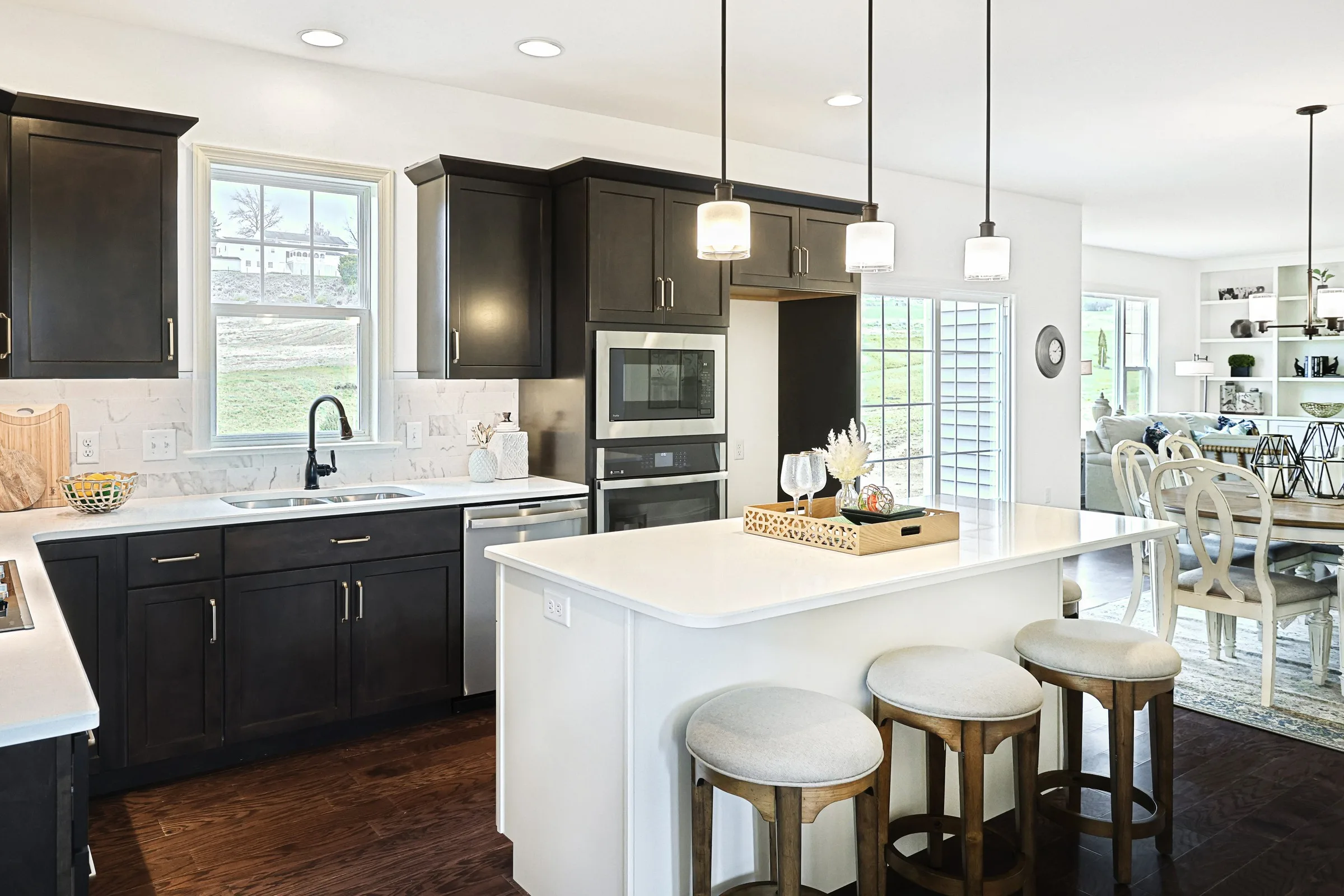 Modern kitchen with dark cabinets, white countertops, island with three stools, hanging pendant lights, built-in oven, and a view into a dining area with a table and chairs. Large windows provide ample natural light.