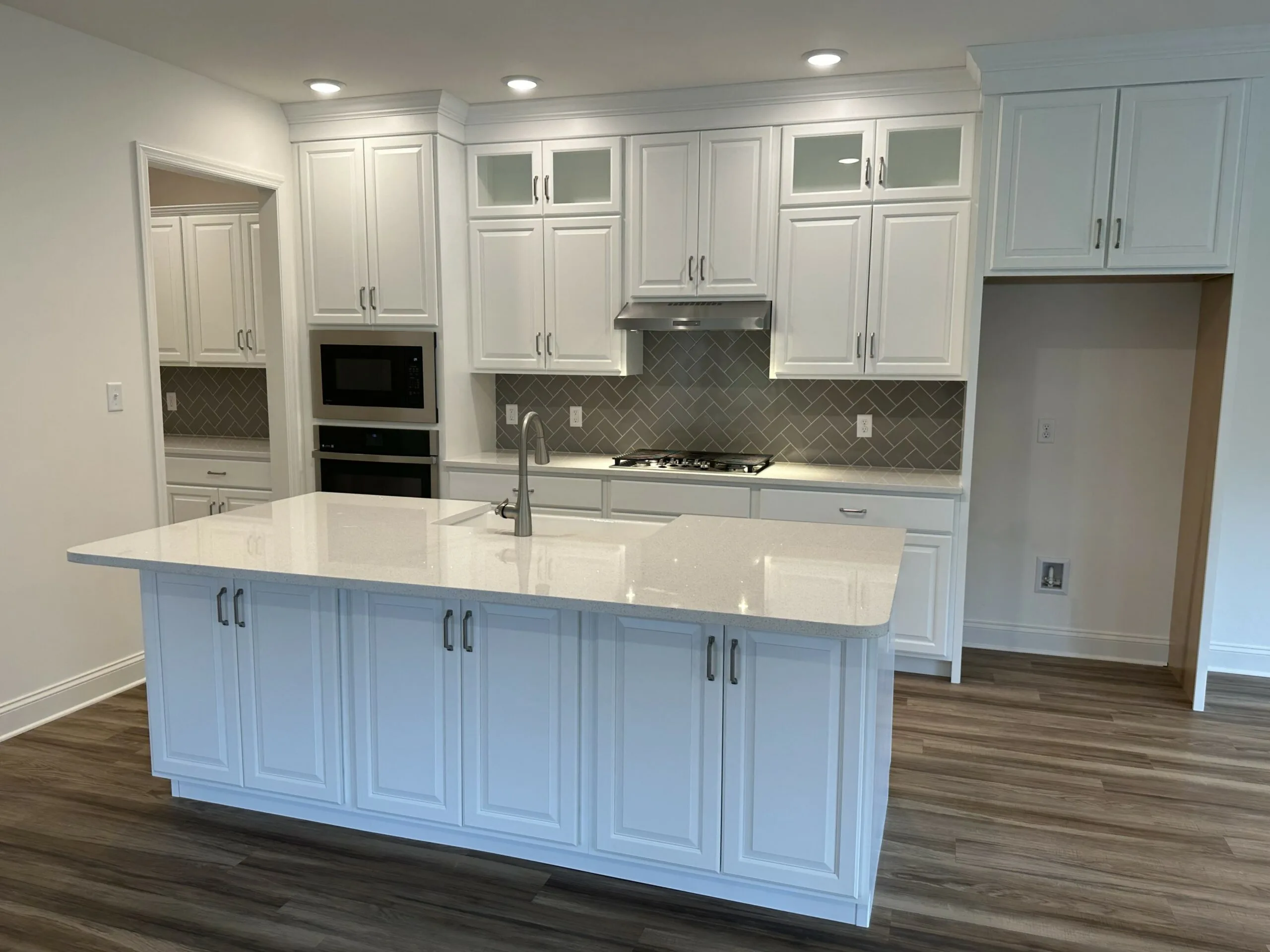 Modern kitchen with white cabinets, a large island with a built-in sink, stainless steel appliances, subway tile backsplash in a herringbone pattern, and wood-look flooring. Recessed lighting brightens the space.