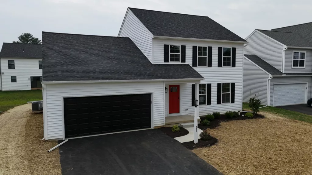 A two-story white house with black shutters, a black garage door, and a red front door. The front yard is partly landscaped with small shrubs and a patch of straw-covered soil beside a paved driveway.