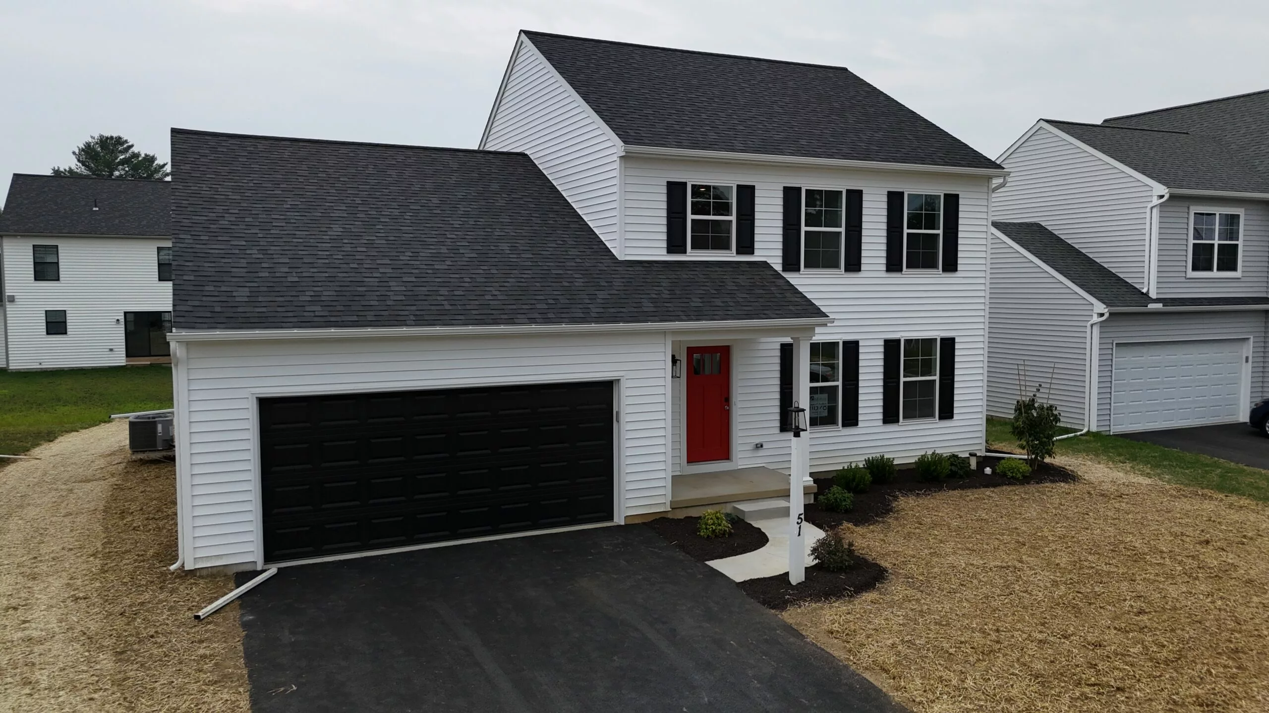 A two-story white house with black shutters, a black garage door, and a red front door. The front yard is partly landscaped with small shrubs and a patch of straw-covered soil beside a paved driveway.
