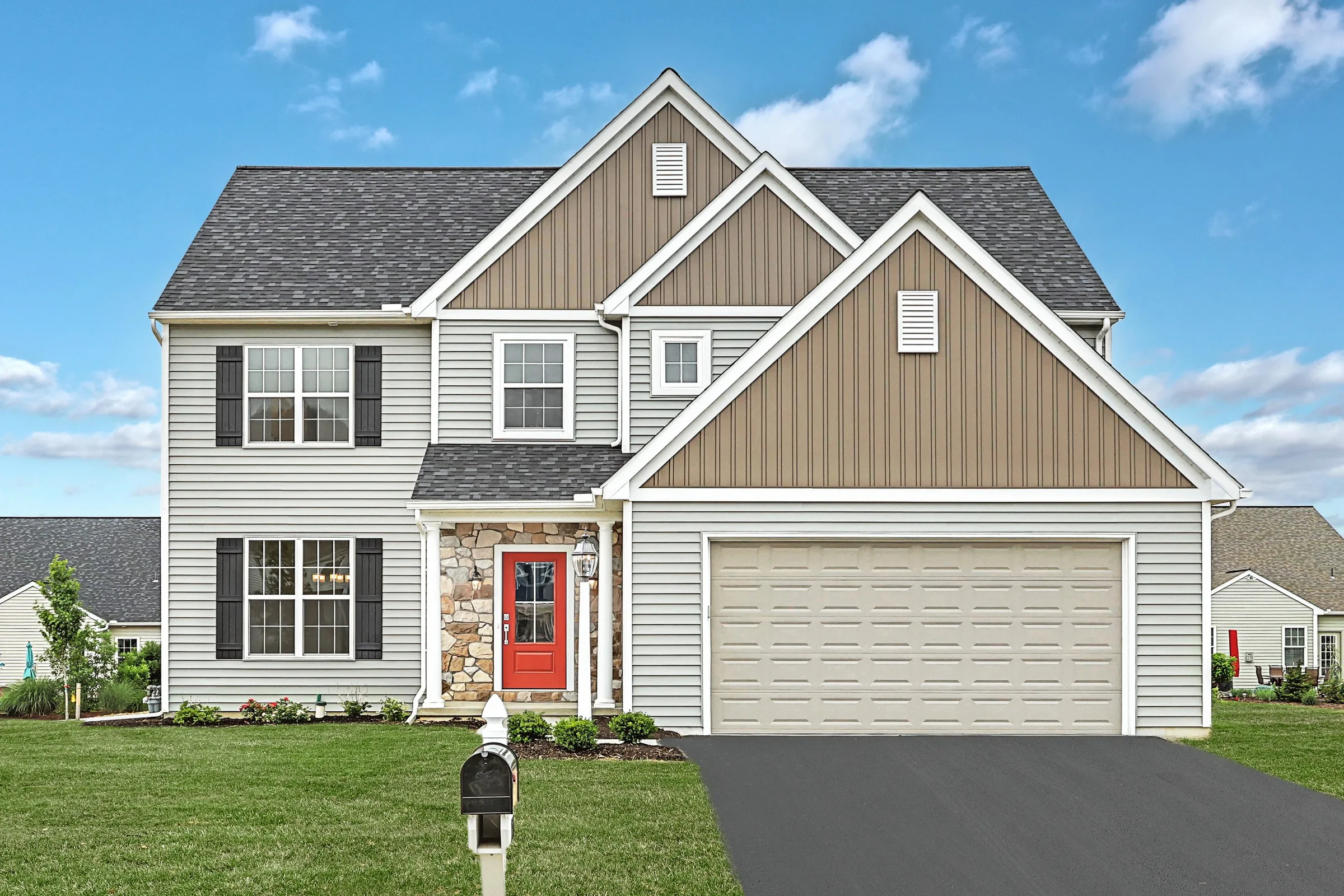 Two-story suburban house with light siding, tan accents, black shutters, a red front door, stone trim, attached two-car garage, and a well-kept lawn under a blue sky with clouds. A mailbox stands by the driveway.