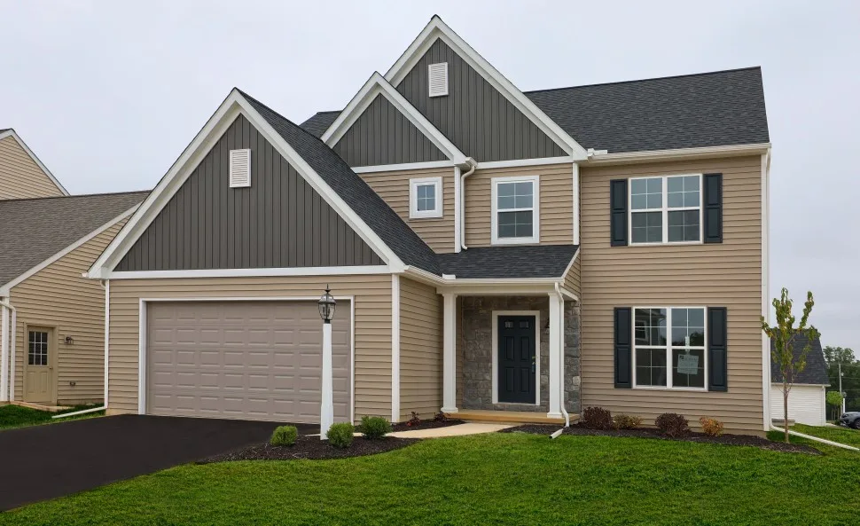 A modern two-story house with beige siding and dark gray accents, featuring a double garage, front porch, and well-maintained lawn with small shrubs and a young tree. The sky is overcast.