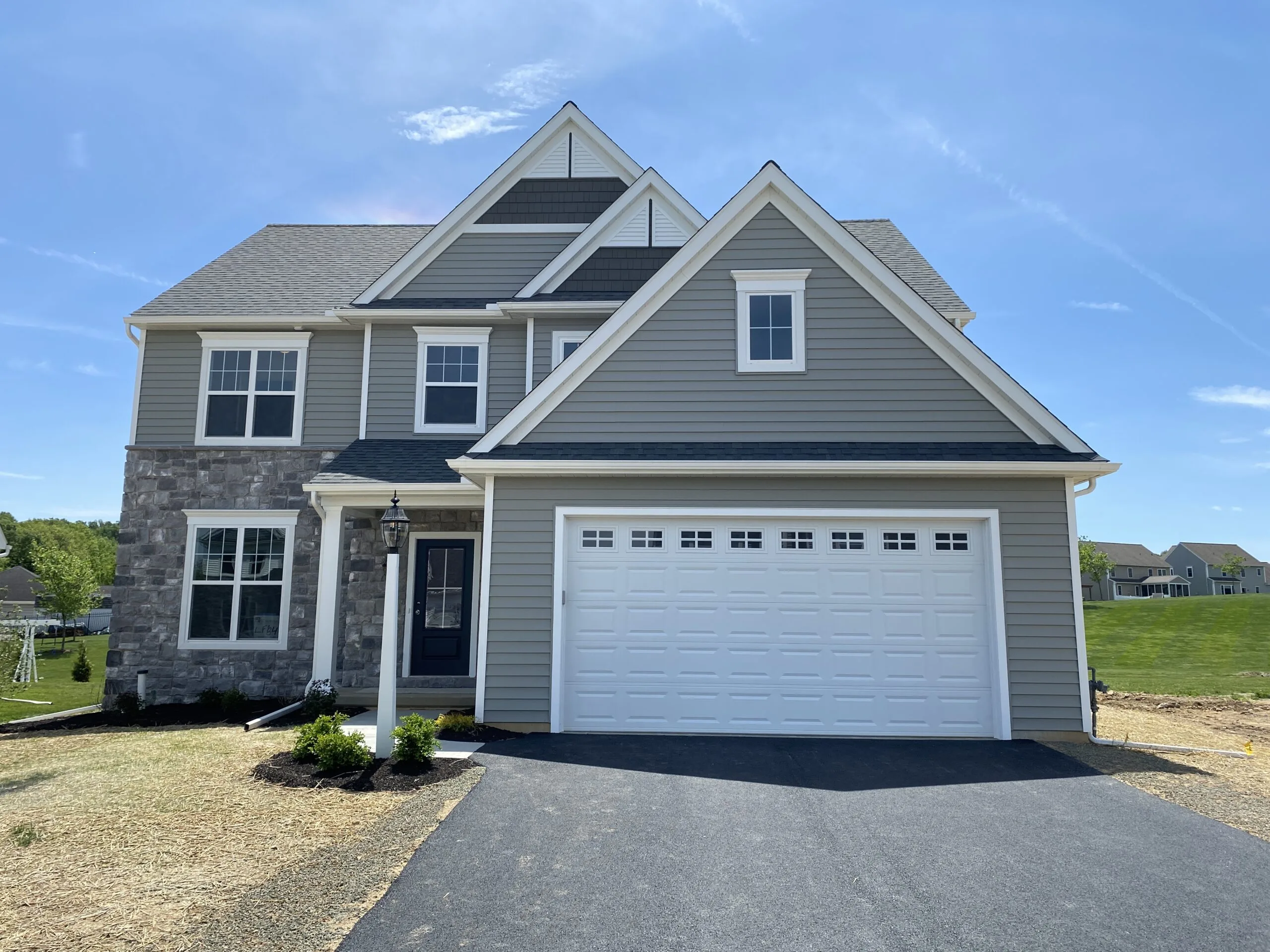 A modern two-story house with gray siding, stone accents, white trim, and a double garage, set on a newly landscaped lot with a blacktop driveway under a clear blue sky.