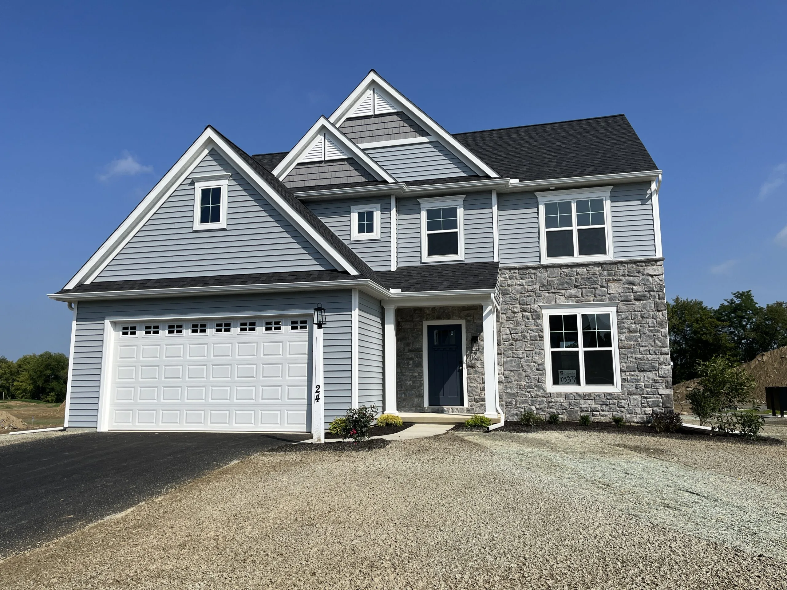 A modern two-story house with light blue siding, a stone facade, white trim, and a two-car garage, situated on a gravel driveway under a clear blue sky.
