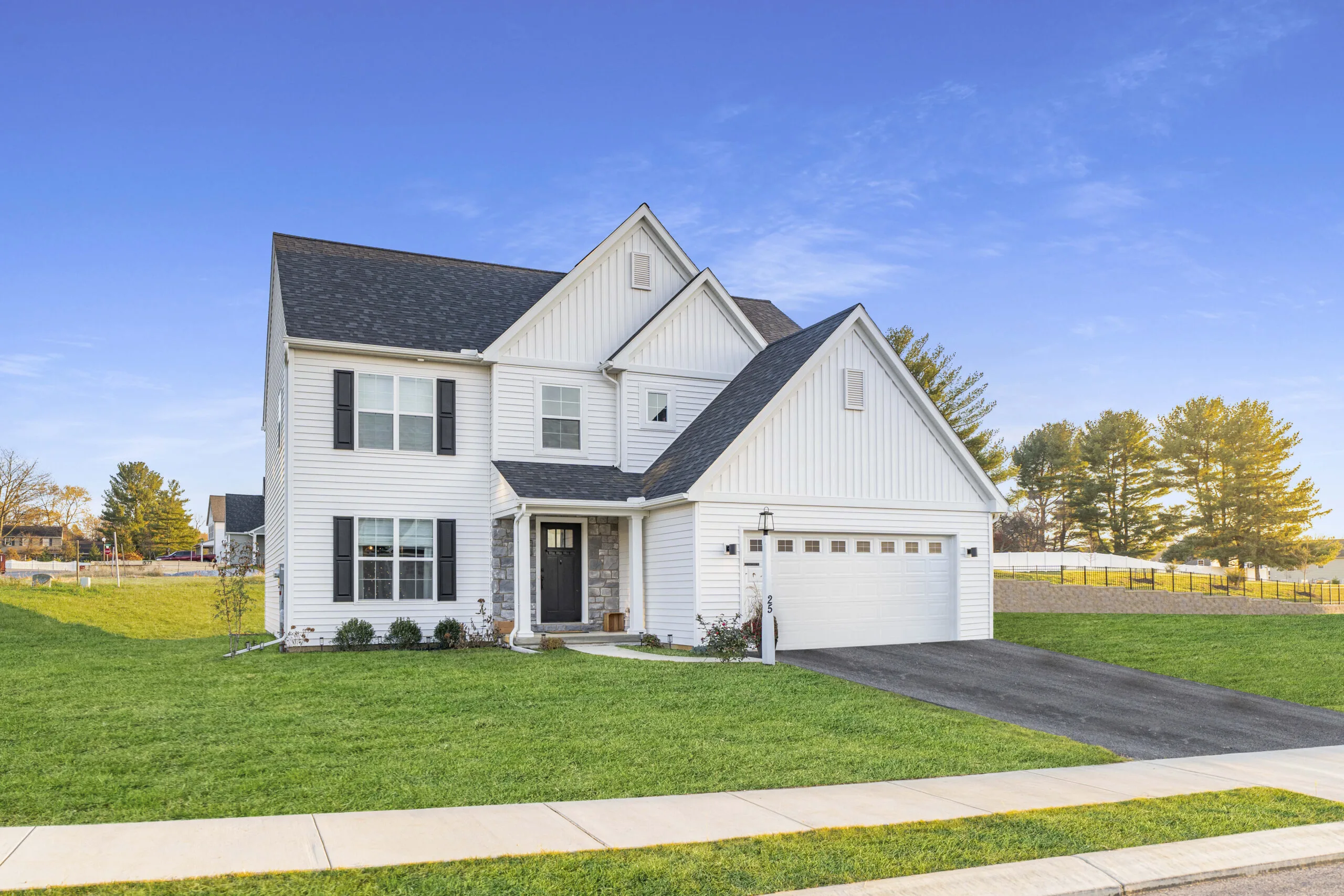Two-story white house with black shutters and a double garage, set on a green lawn with a paved driveway, trees, and a clear blue sky in the background.