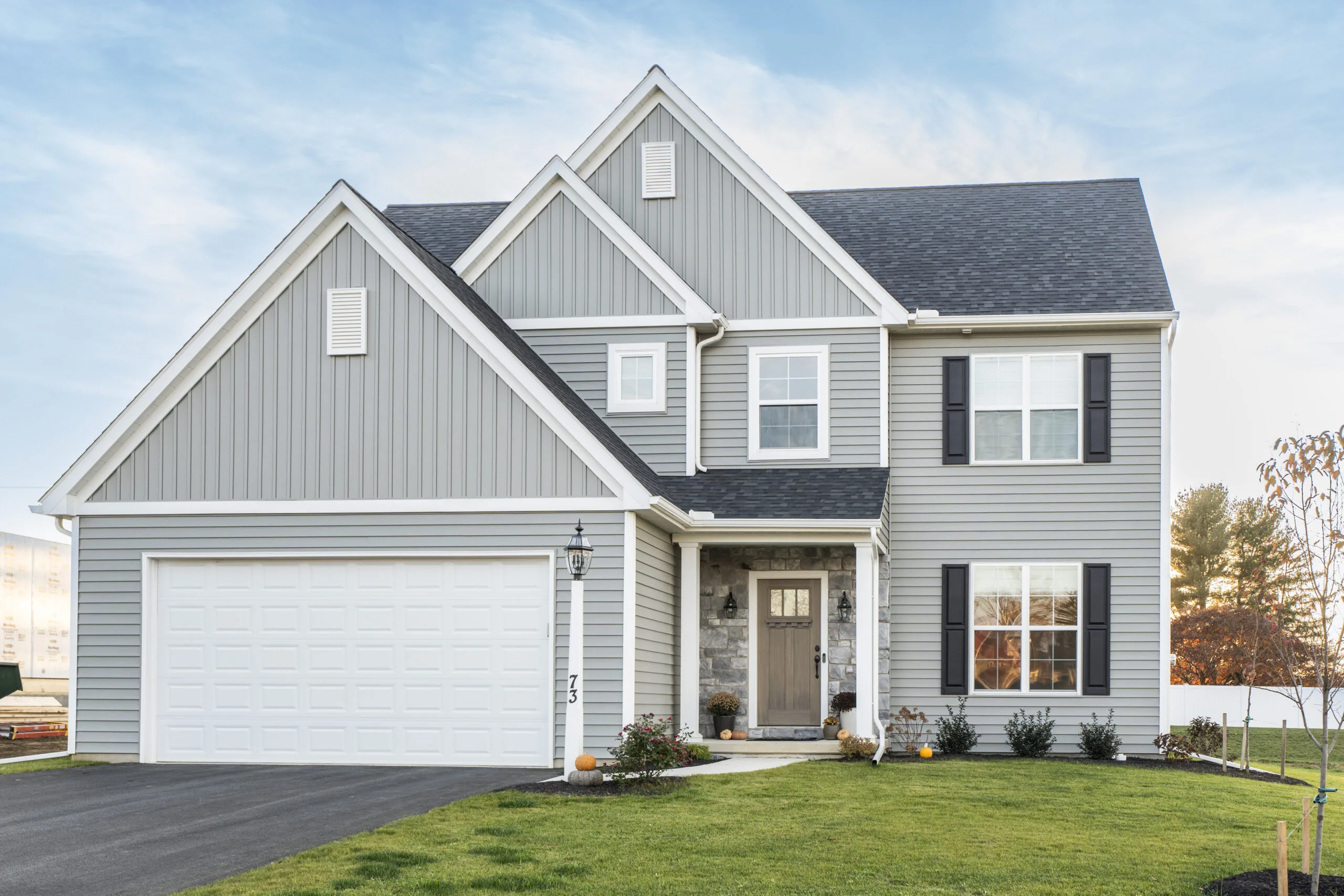 A modern two-story gray house with white trim, a two-car garage, and a neatly maintained lawn. The house has large front windows, dark shutters, and a paved driveway under a blue sky.