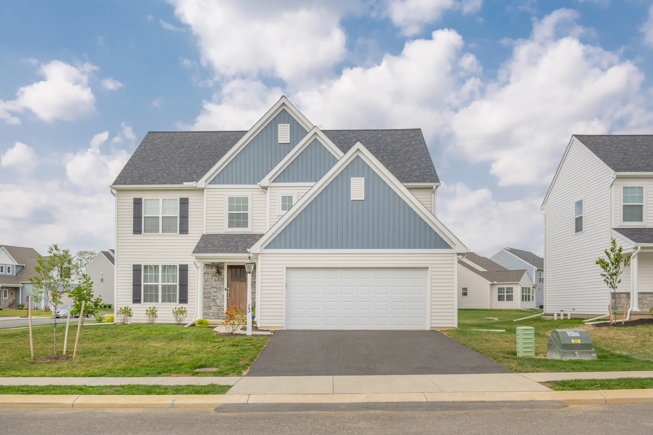 A modern two-story suburban house with gray and white siding, a double garage, a paved driveway, and a neatly trimmed lawn under a partly cloudy sky.