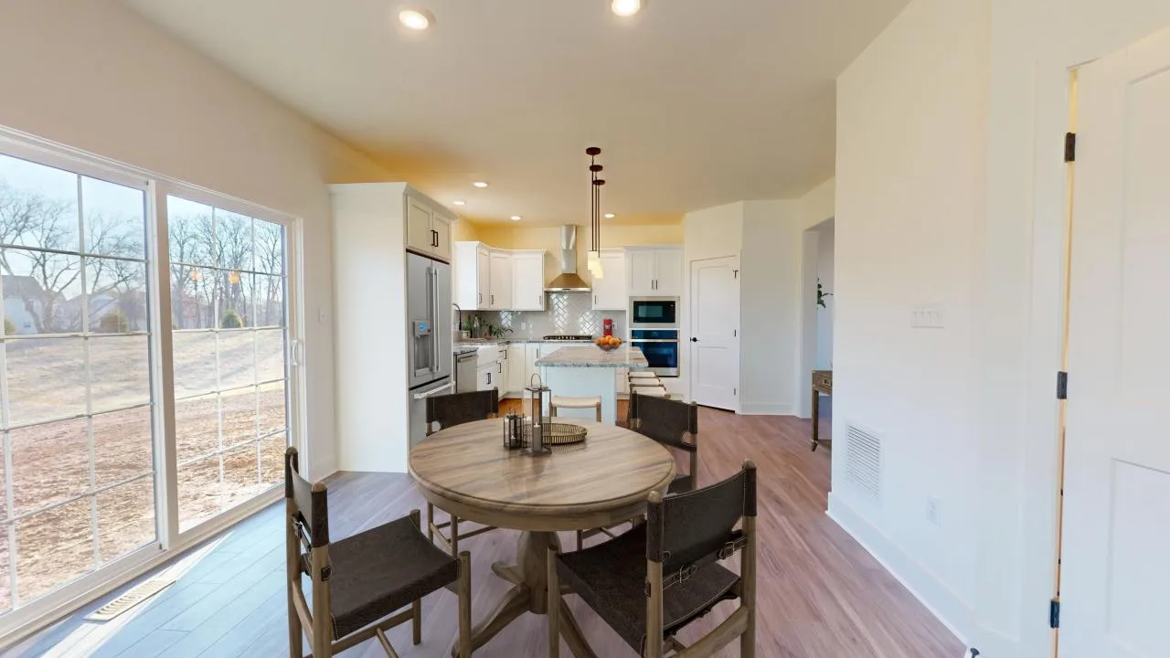A modern kitchen and dining area with a round wooden table and four chairs, light wood flooring, white cabinets, stainless steel appliances, and large sliding glass doors letting in natural light.