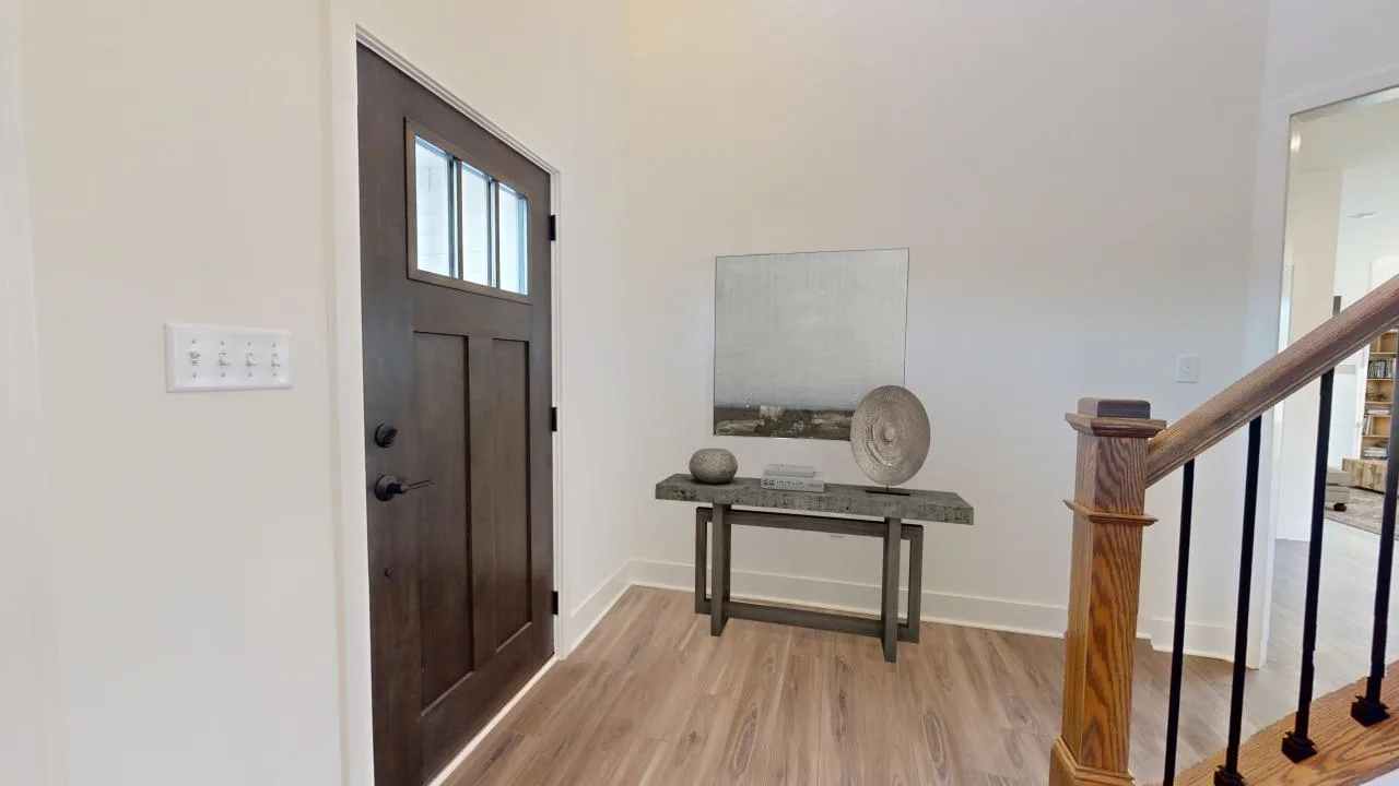 A modern entryway with a dark wooden front door, a small console table holding decorative objects and artwork, light wood flooring, and a staircase with a wooden handrail.