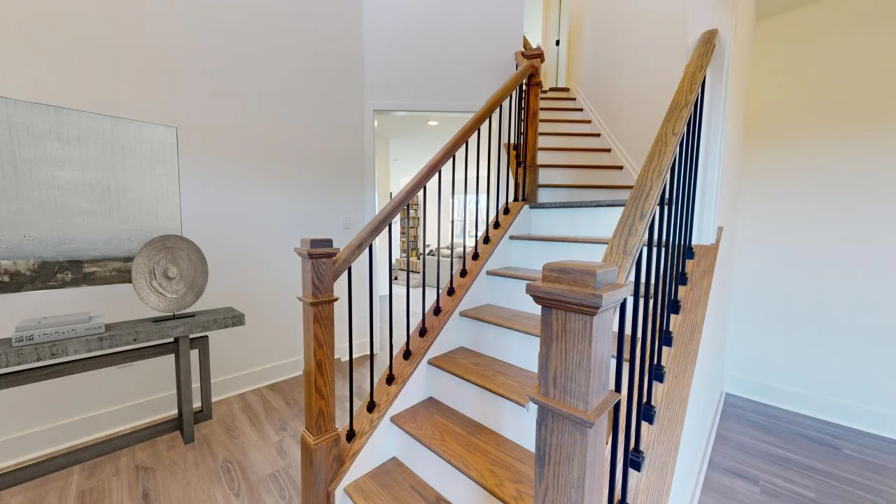 A wooden staircase with black iron balusters and light-colored steps ascends in a well-lit, modern hallway. A decorative console table with art pieces sits against the white wall on the left.