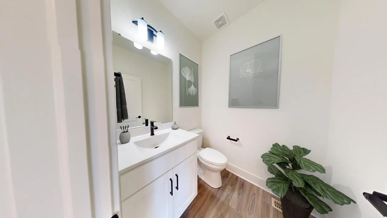 A modern bathroom with white walls, a white vanity and sink, a large mirror, a toilet, wooden flooring, minimal decor, two floral artworks, and a green potted plant in the corner.