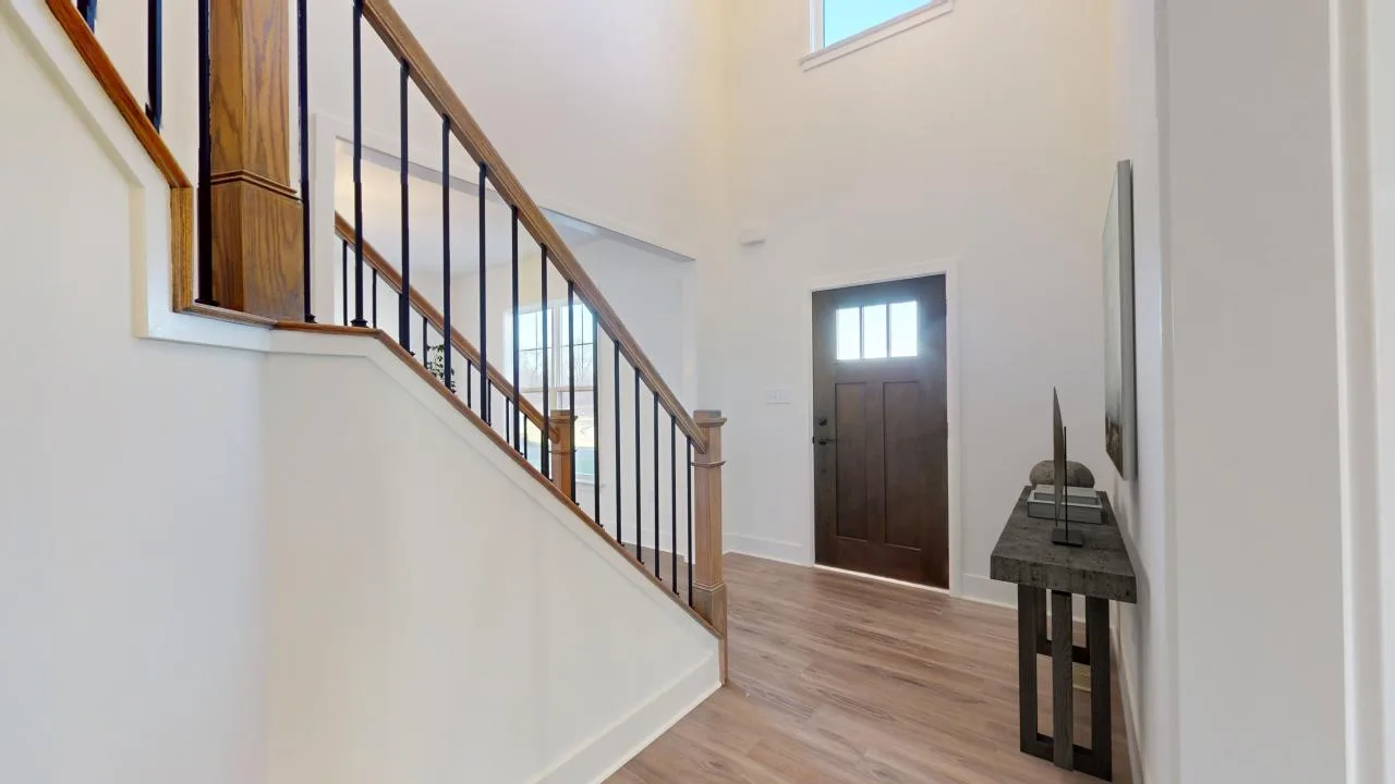 A bright, modern entryway with light wood floors, white walls, a dark wooden front door with glass panels, a simple black console table, and a staircase with wooden handrails and black spindles.