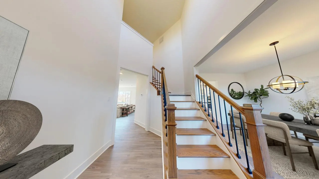A modern home entryway with wooden stairs and black railings, leading to an open dining area with a round black chandelier, light walls, and hardwood floors. Bright natural light fills the space.